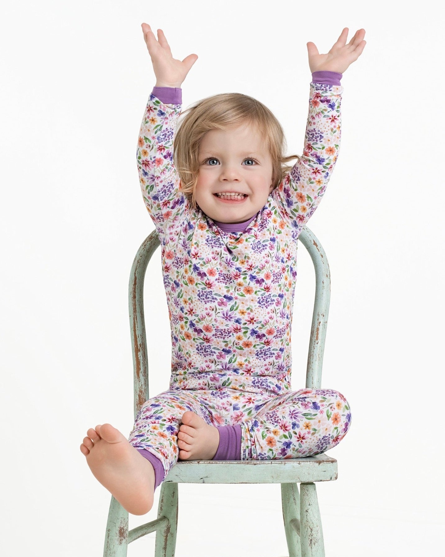 A young girl in floral pajamas sits on a chair with her arms raised.