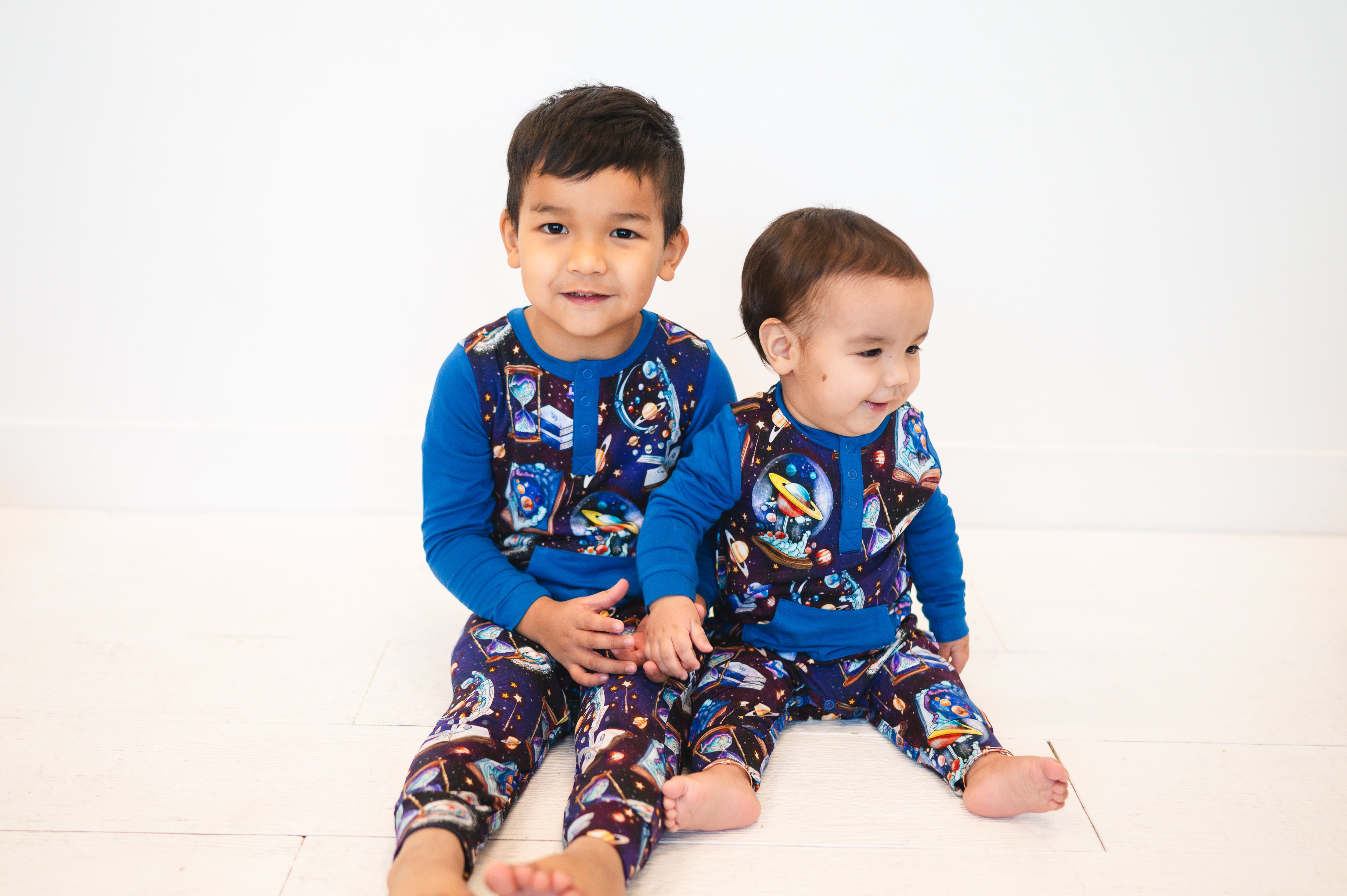 Two young boys in blue space-themed bamboo rompers sit on a white floor.