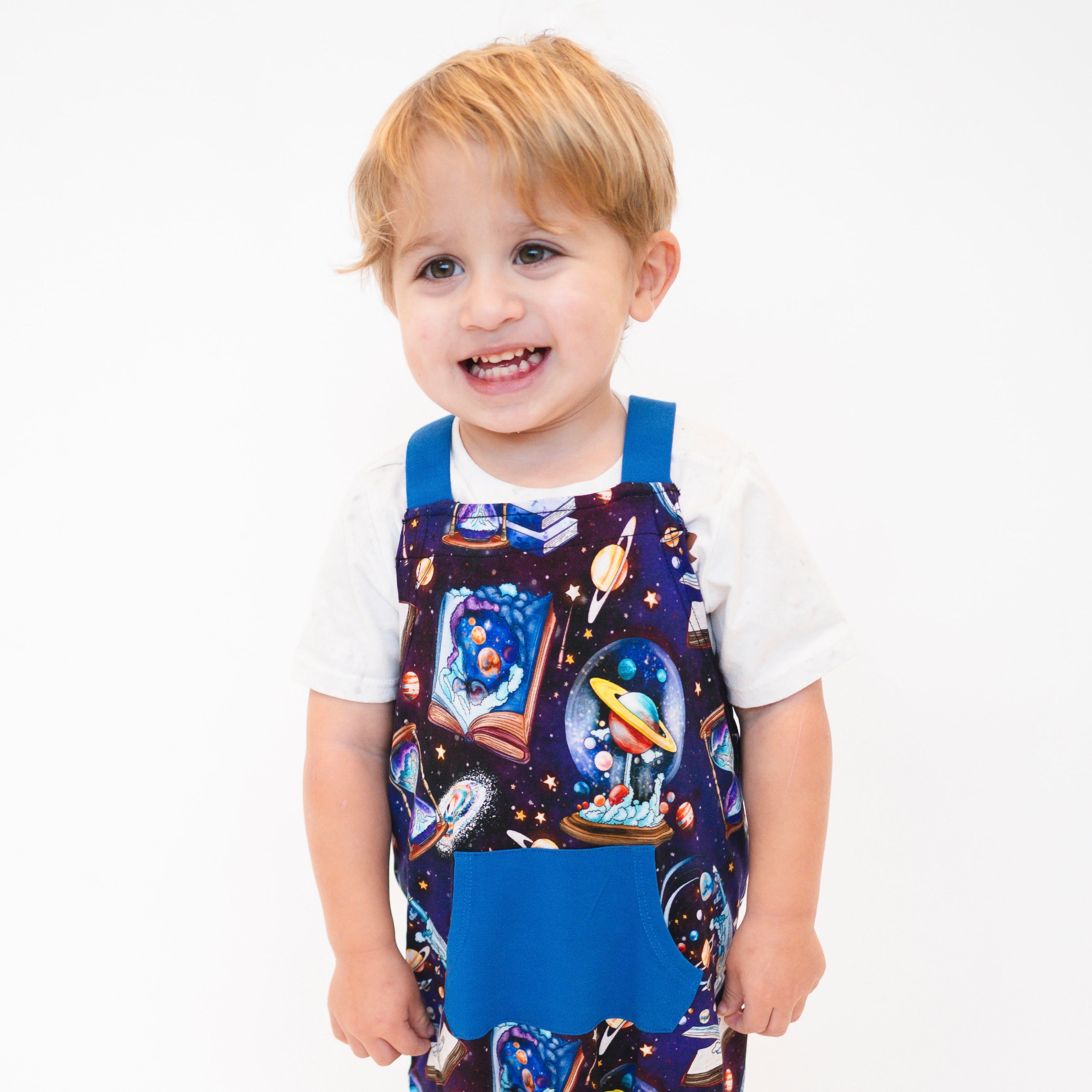 Toddler in white shirt and navy overalls with a space print and blue pocket.