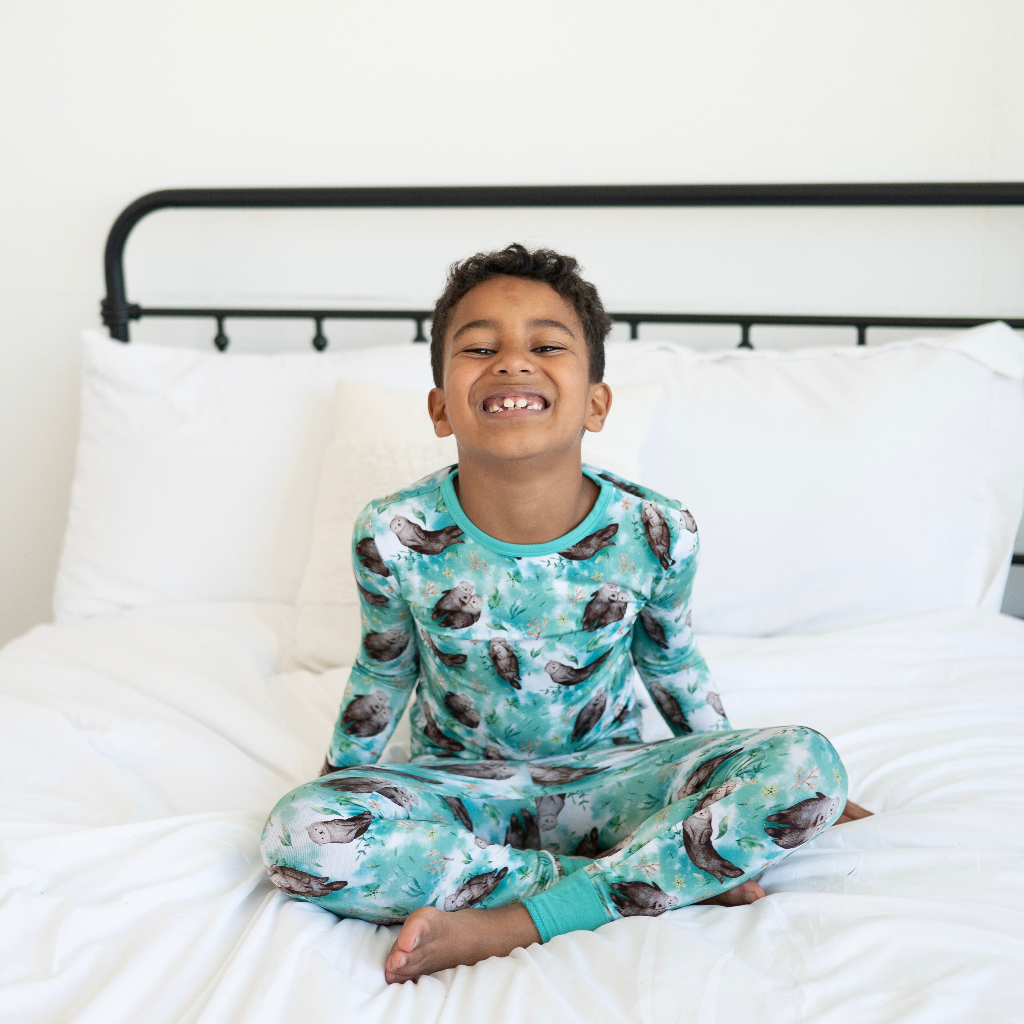 A young boy smiles while wearing otter-print pajamas on a white bed.