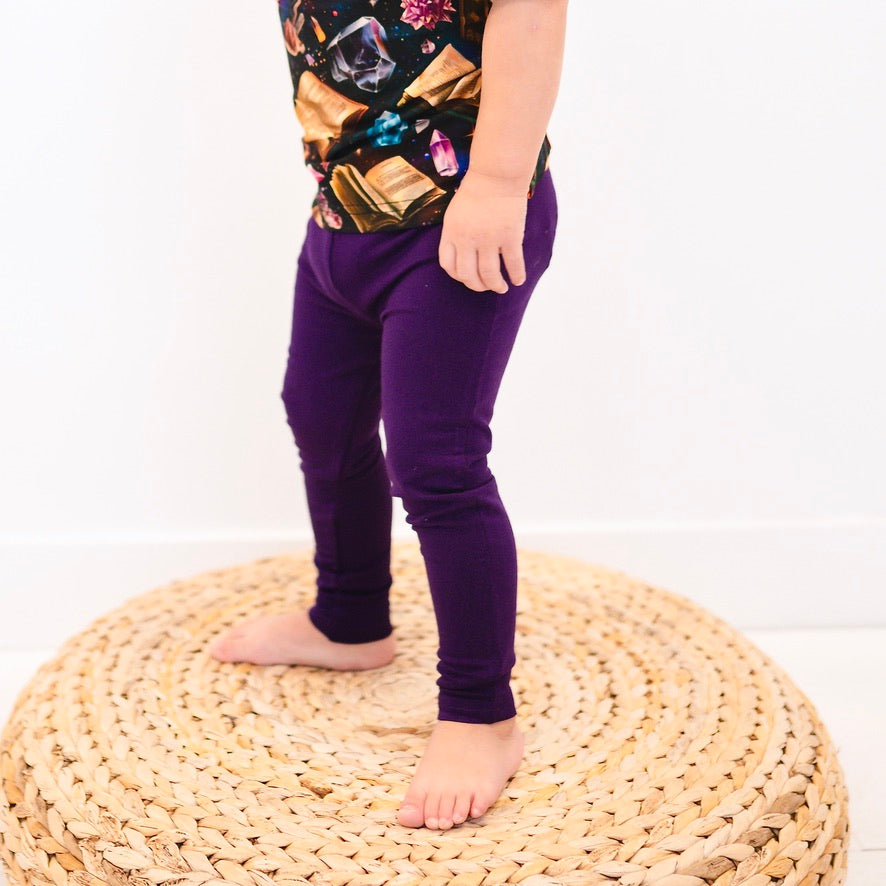 A child wears purple bamboo leggings and a patterned shirt, standing on a woven stool.
