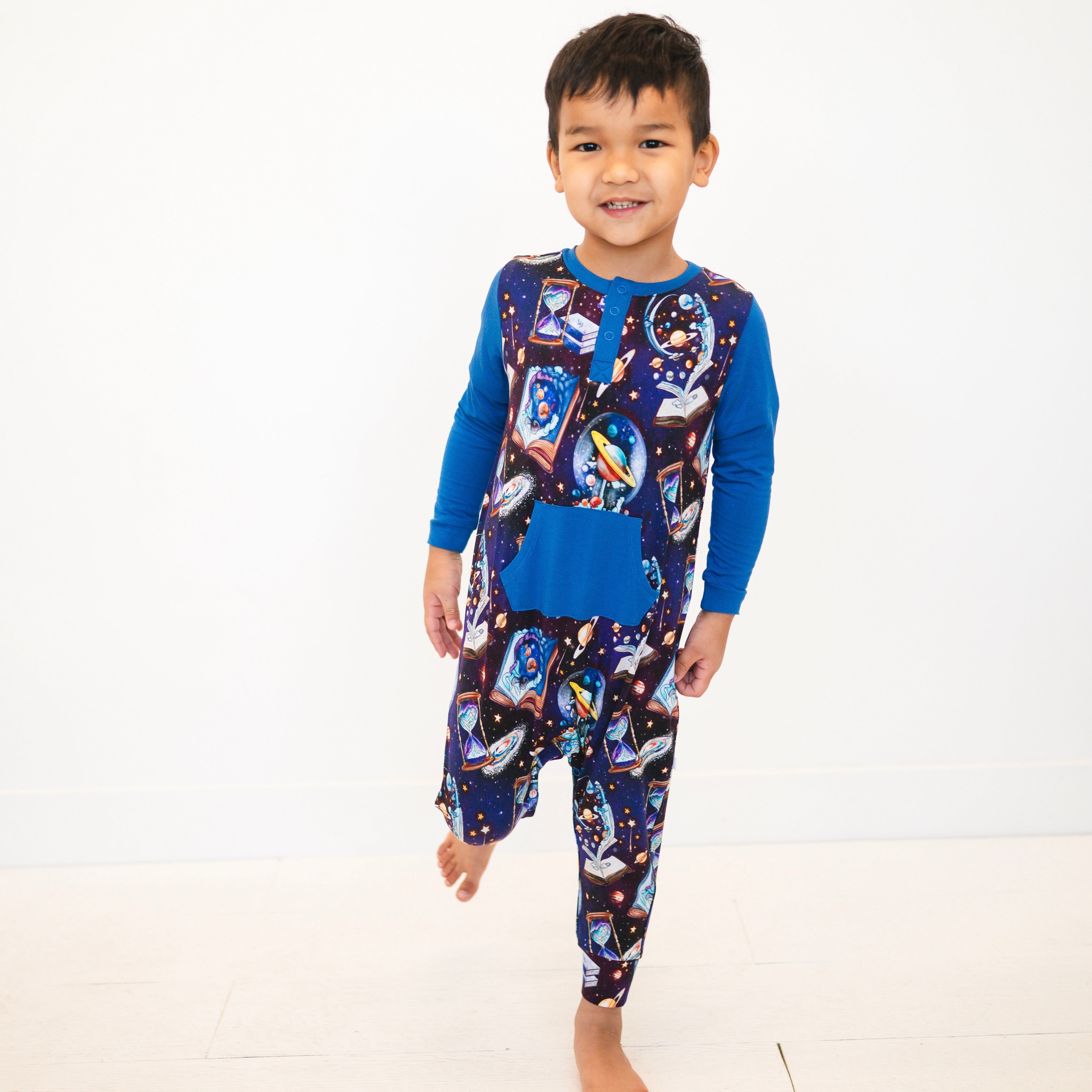 A young boy stands on one leg wearing a blue and space-themed romper.