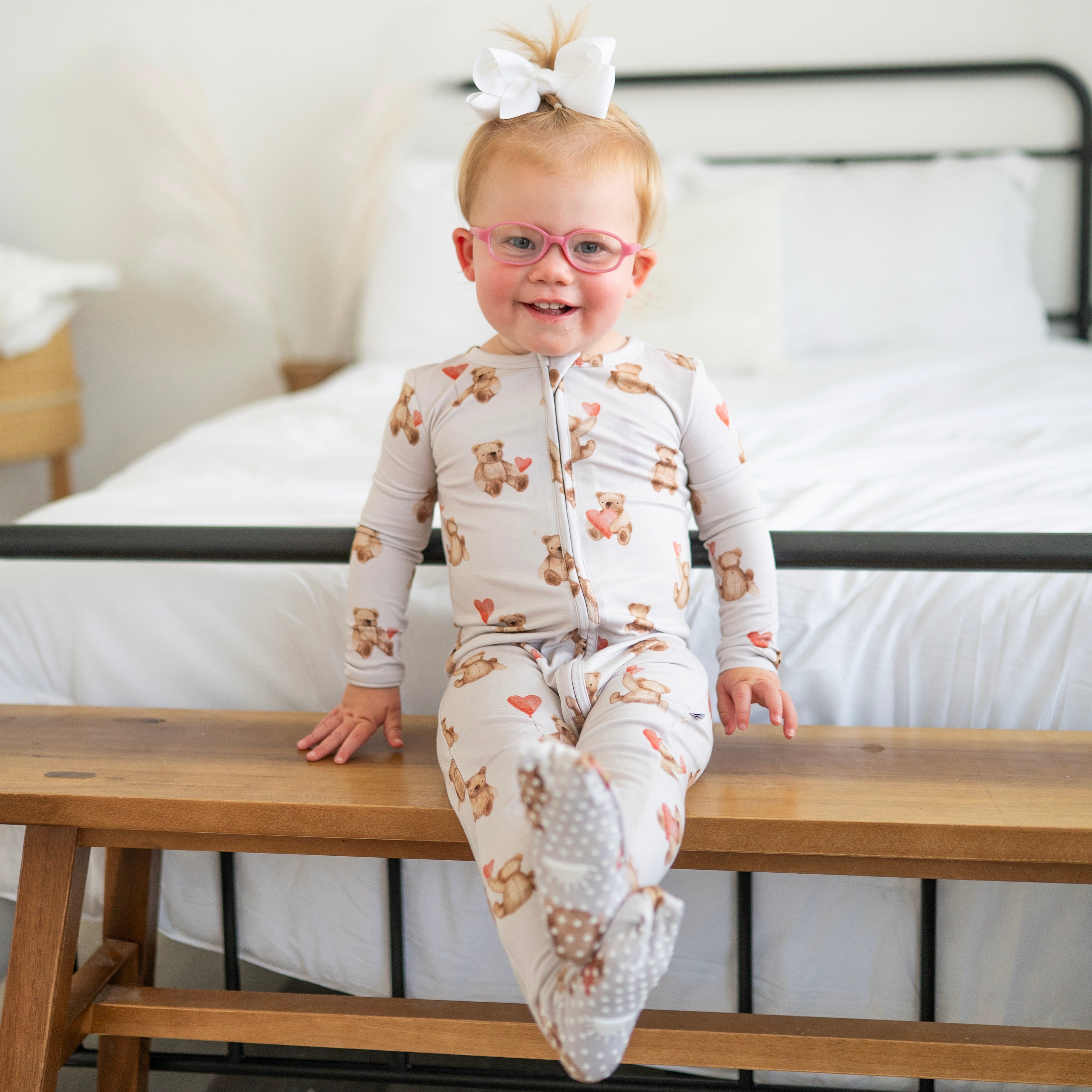 A baby girl wearing a teddy bear print footie pajamas and pink glasses sits on a wooden bench.