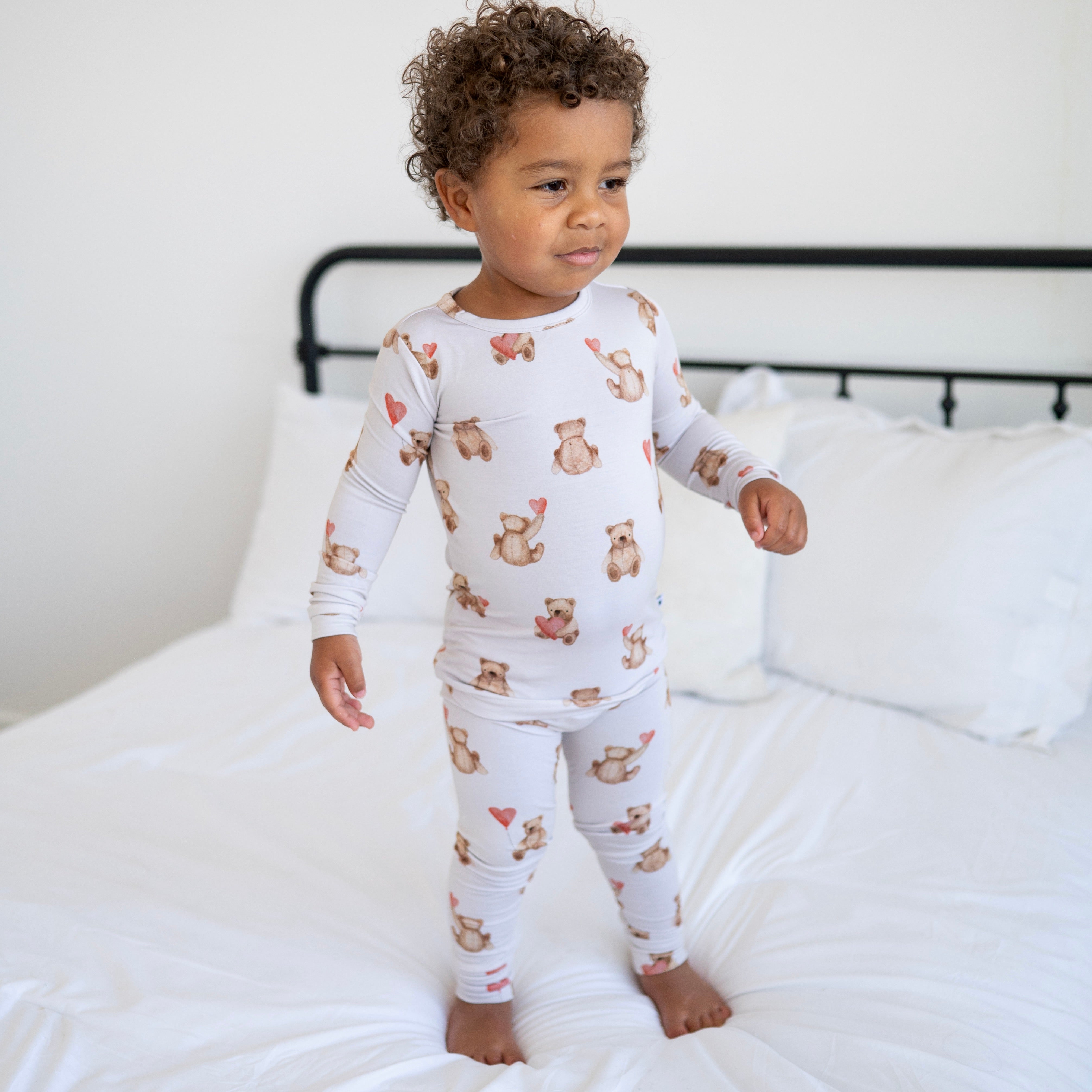 A young child stands on a bed wearing a light-colored pajama set with a teddy bear and heart print.