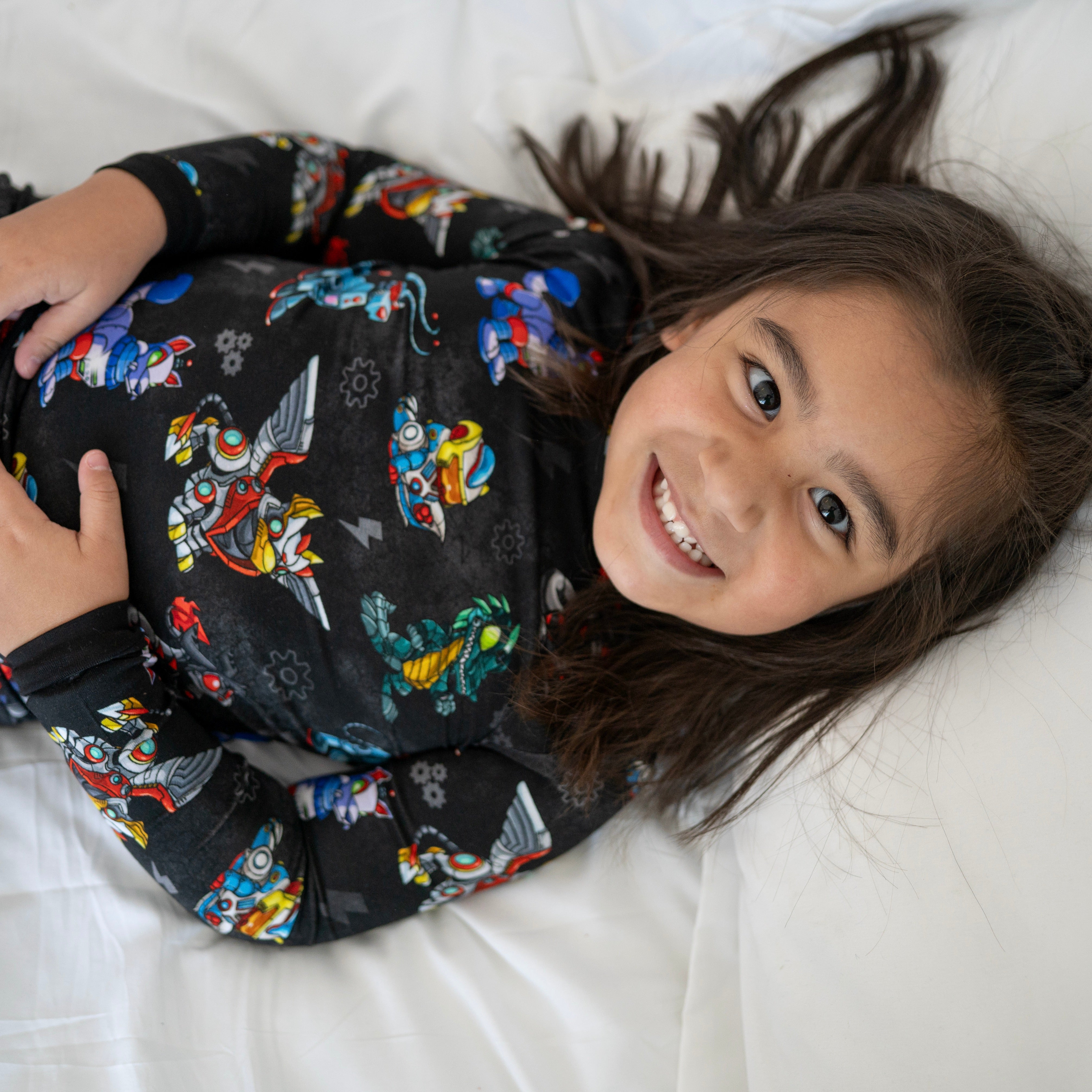 A child in black pajamas with a robot print smiles on a white bed.