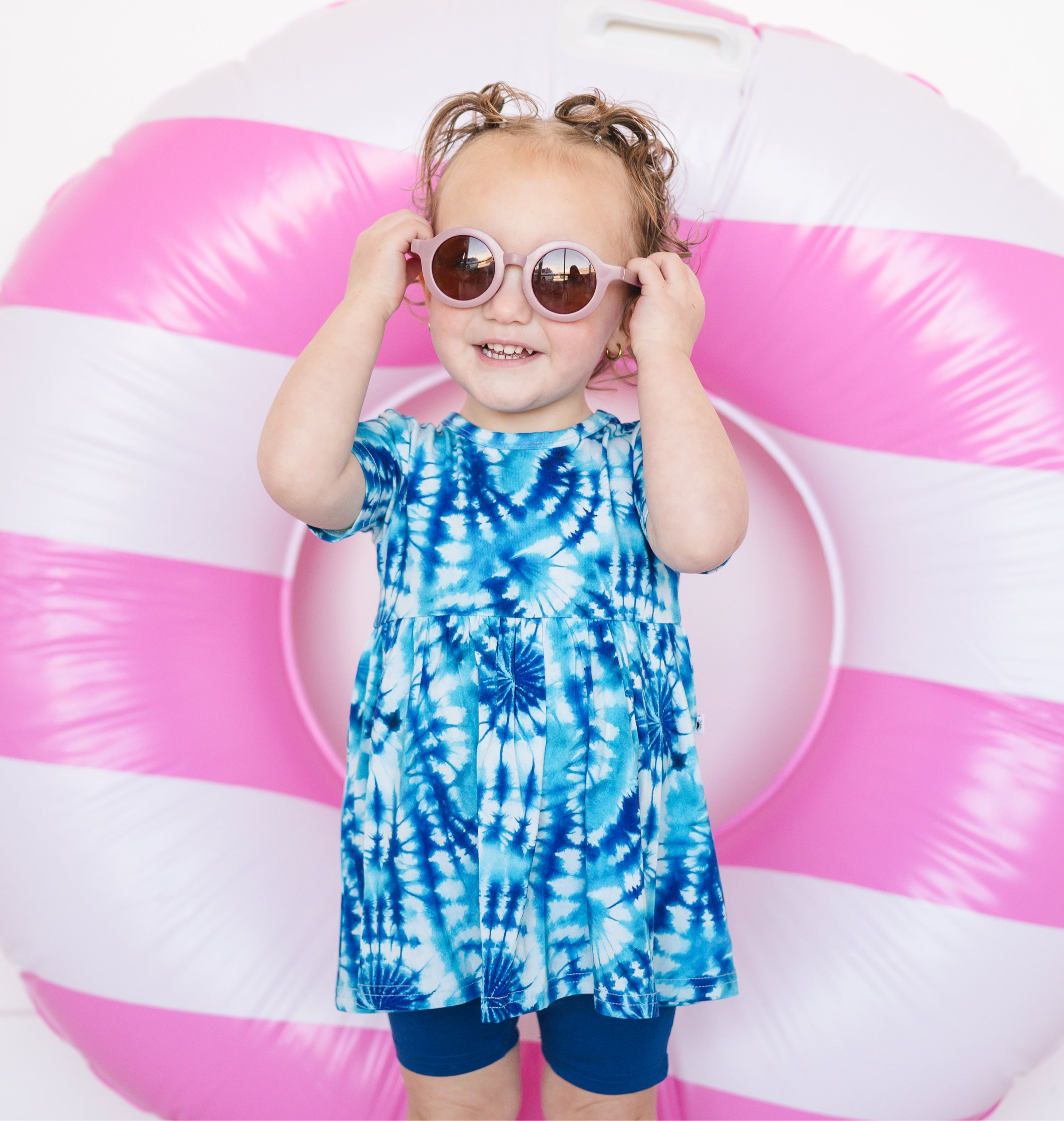 A young girl wears a blue tie-dye peplum top and blue shorts, holding pink sunglasses in front of a pink and white inflatable ring.