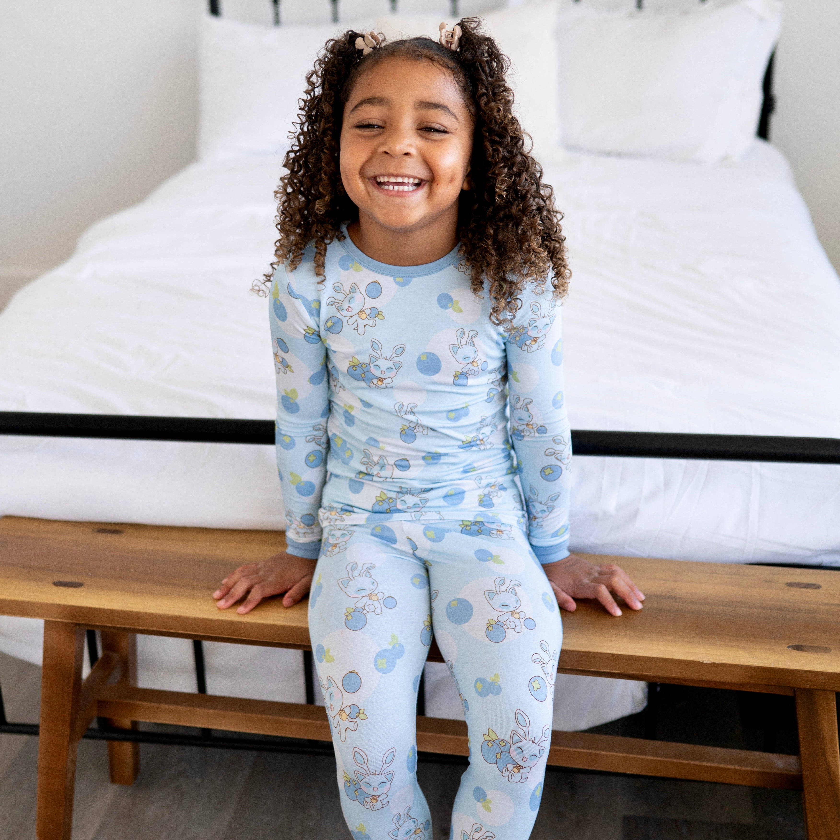 A young girl in light blue pajamas with a blueberry and baby Aisha print sits on a wooden bench.