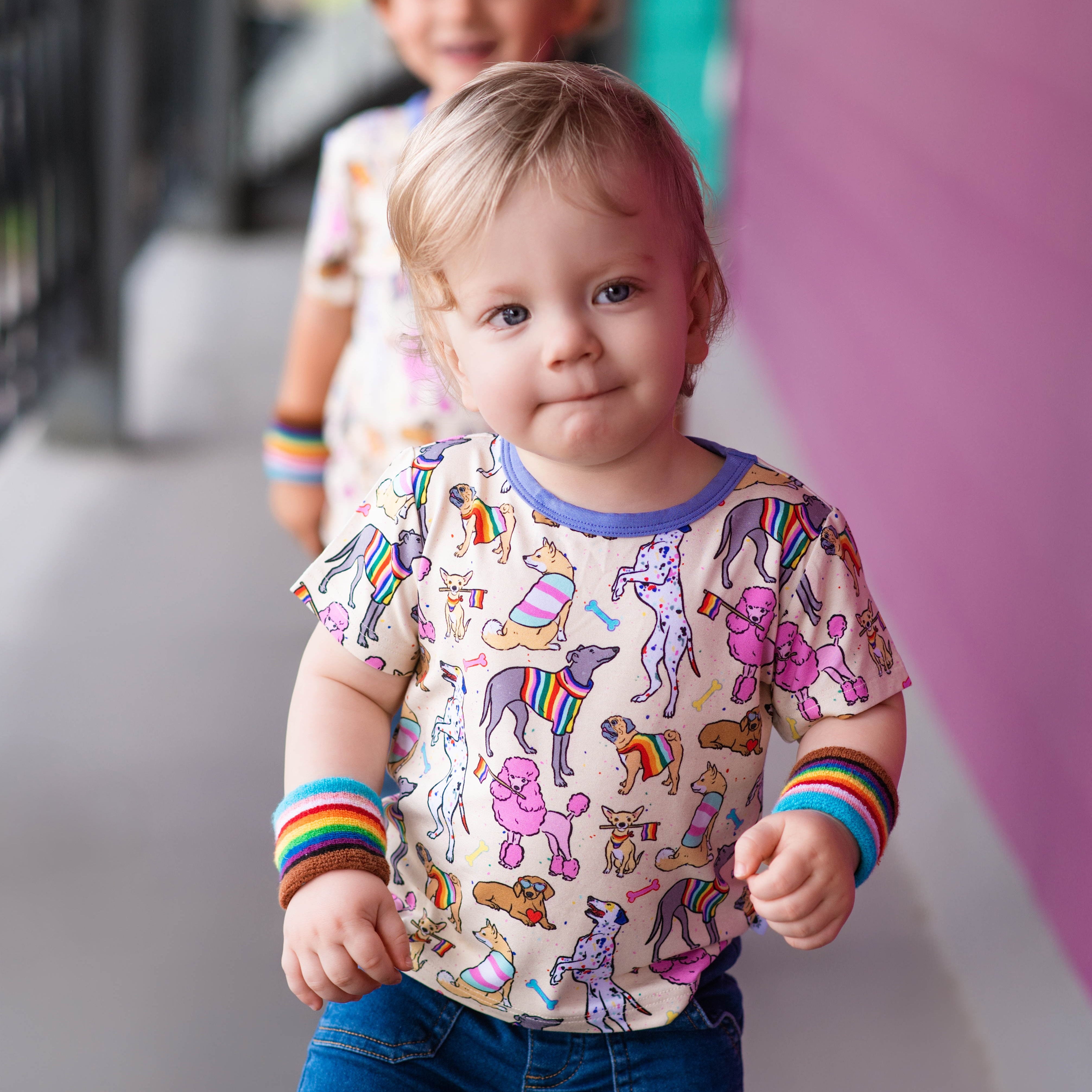 Toddler wearing a light-colored t-shirt with a print of dogs in rainbow sweaters and holding rainbow flags.