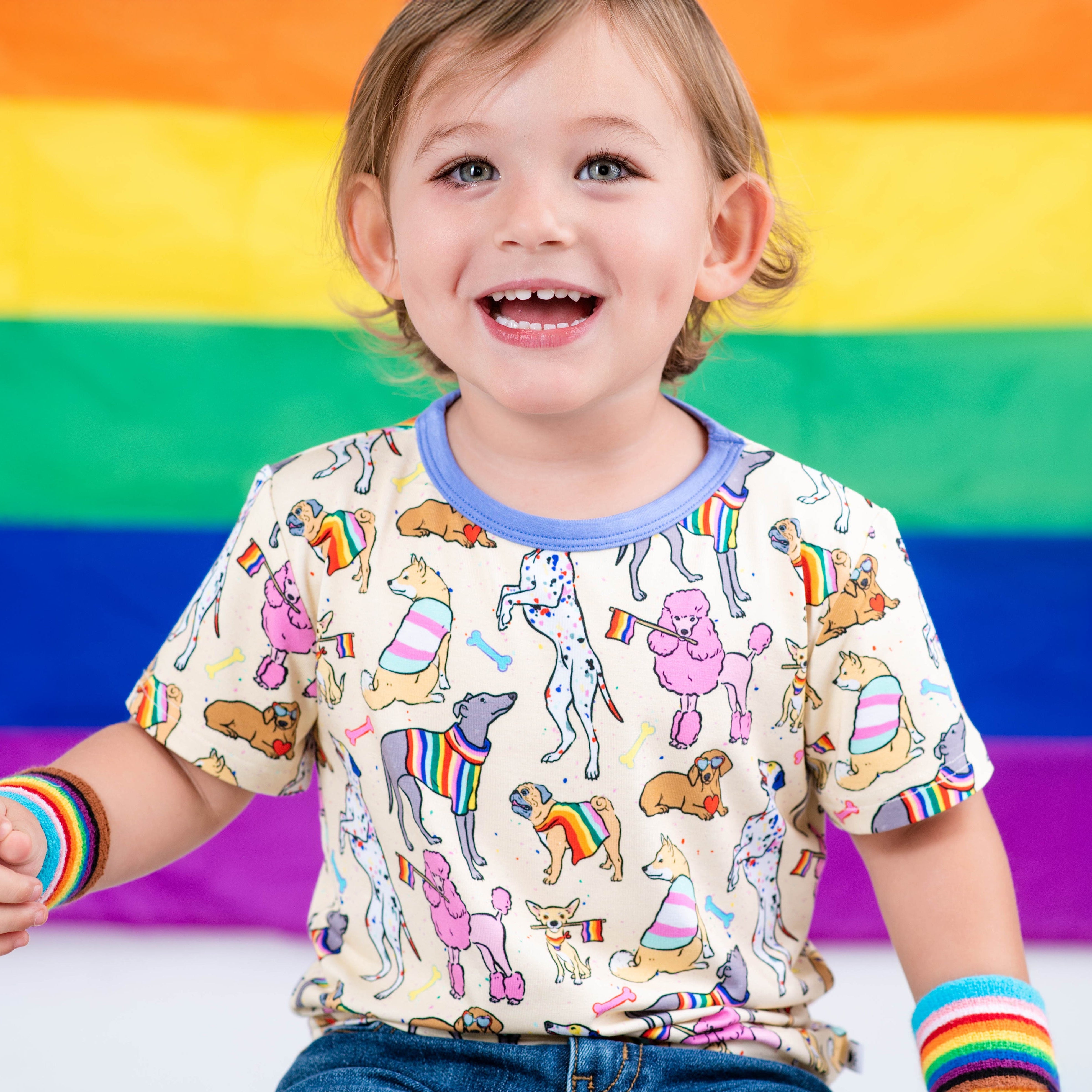 Toddler wearing a light-colored t-shirt with a print of various dogs, some wearing rainbow stripes, in front of a rainbow flag.