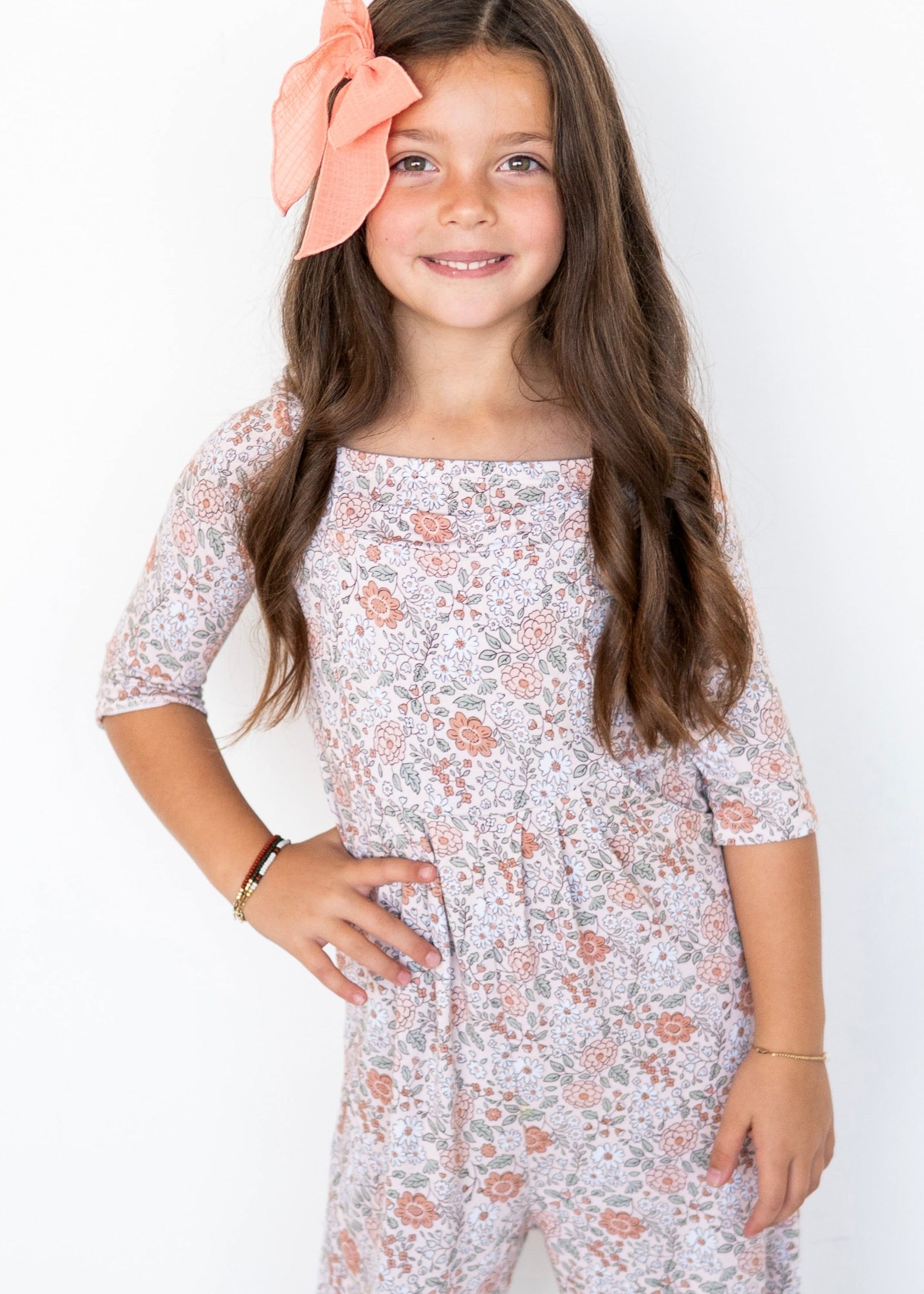 A young girl wears a floral print romper with a coral bow in her hair.