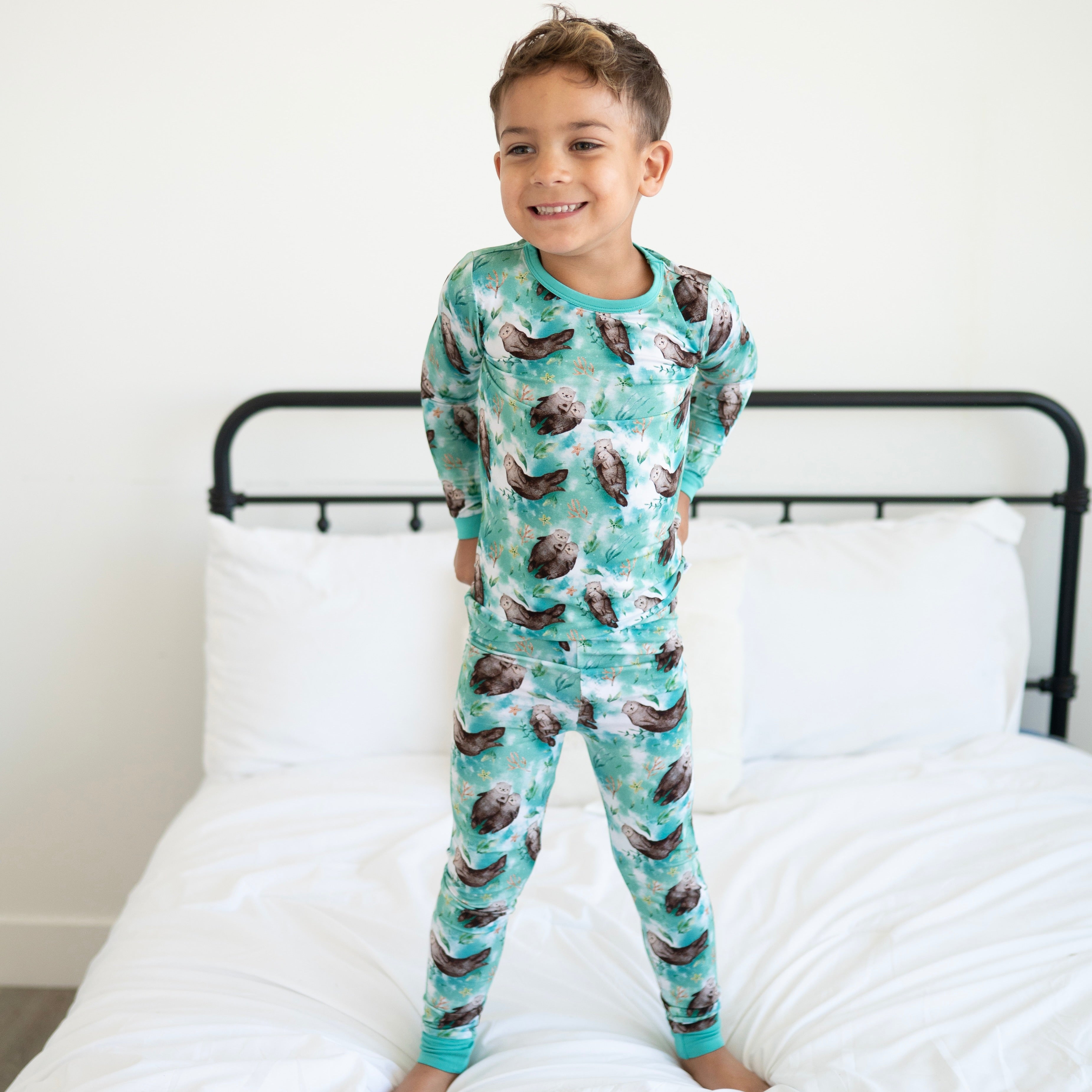 A young boy stands on a bed wearing a long-sleeved pajama set with an otter print.