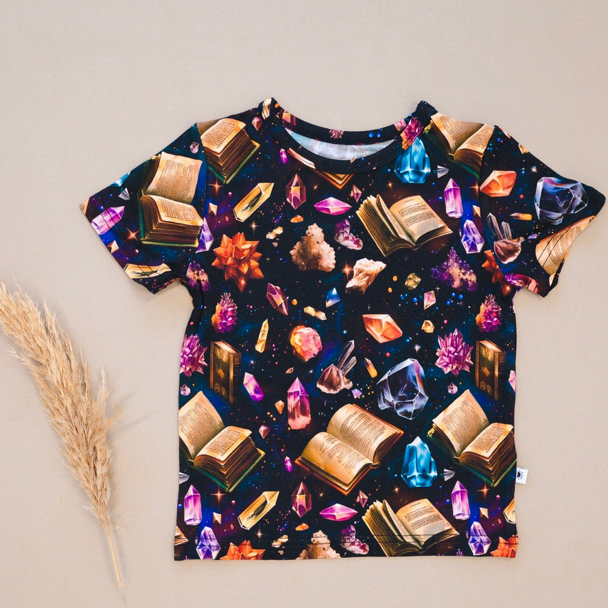 T-shirt with a dark background featuring an all-over print of open books and colorful crystals.