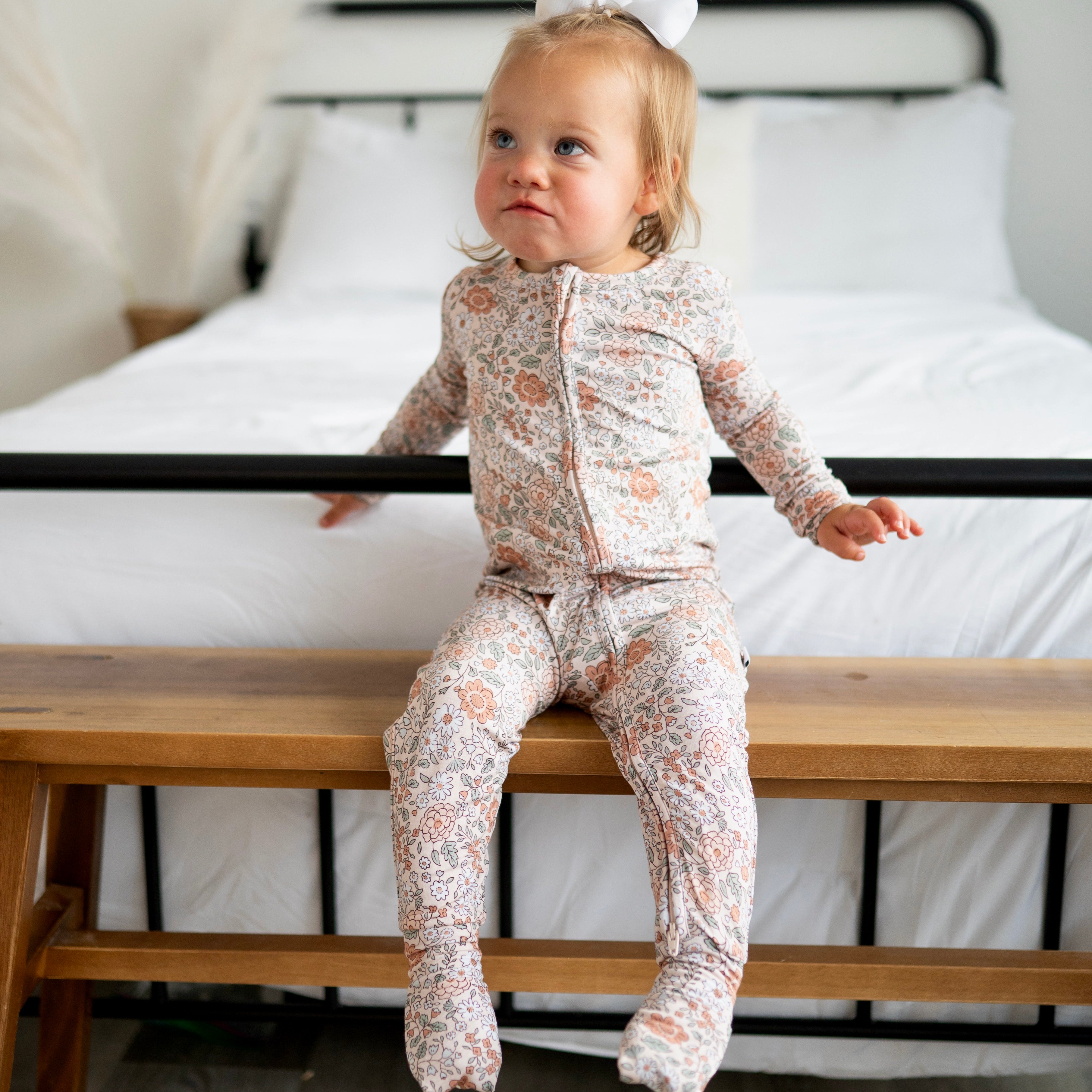 A toddler girl wearing a vintage floral zip-up pajama set sits on a wooden bench.