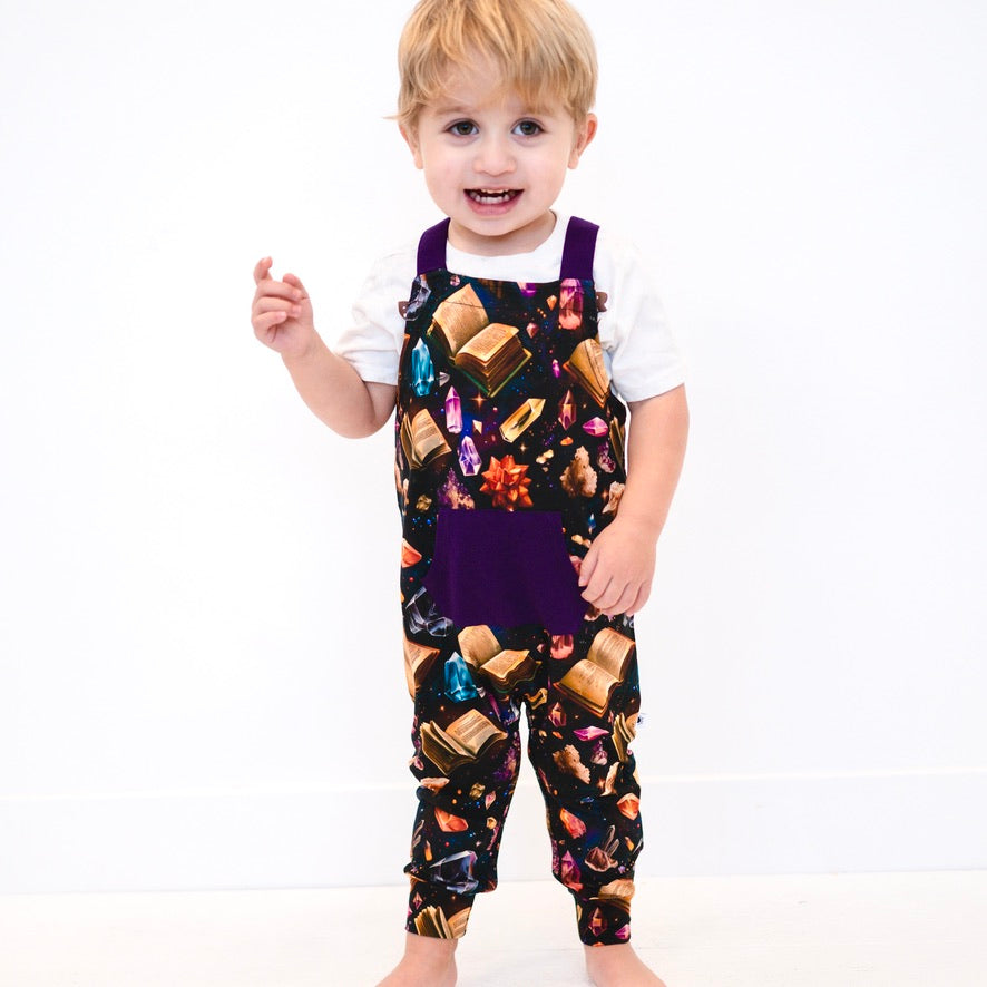 Toddler overalls with a crystal and book print and purple pocket.