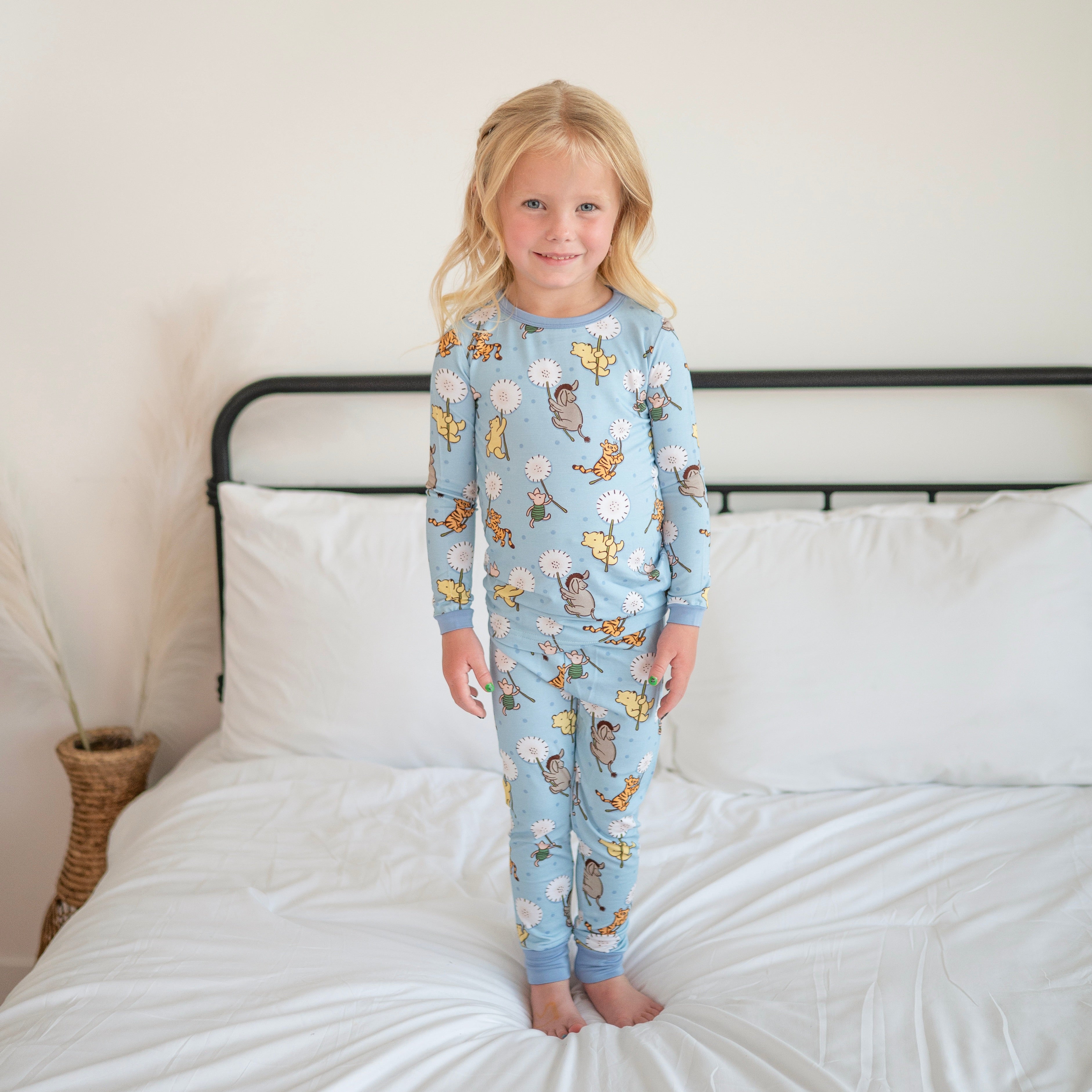 A young girl stands on a bed wearing blue pajamas with a Winnie the Pooh print.