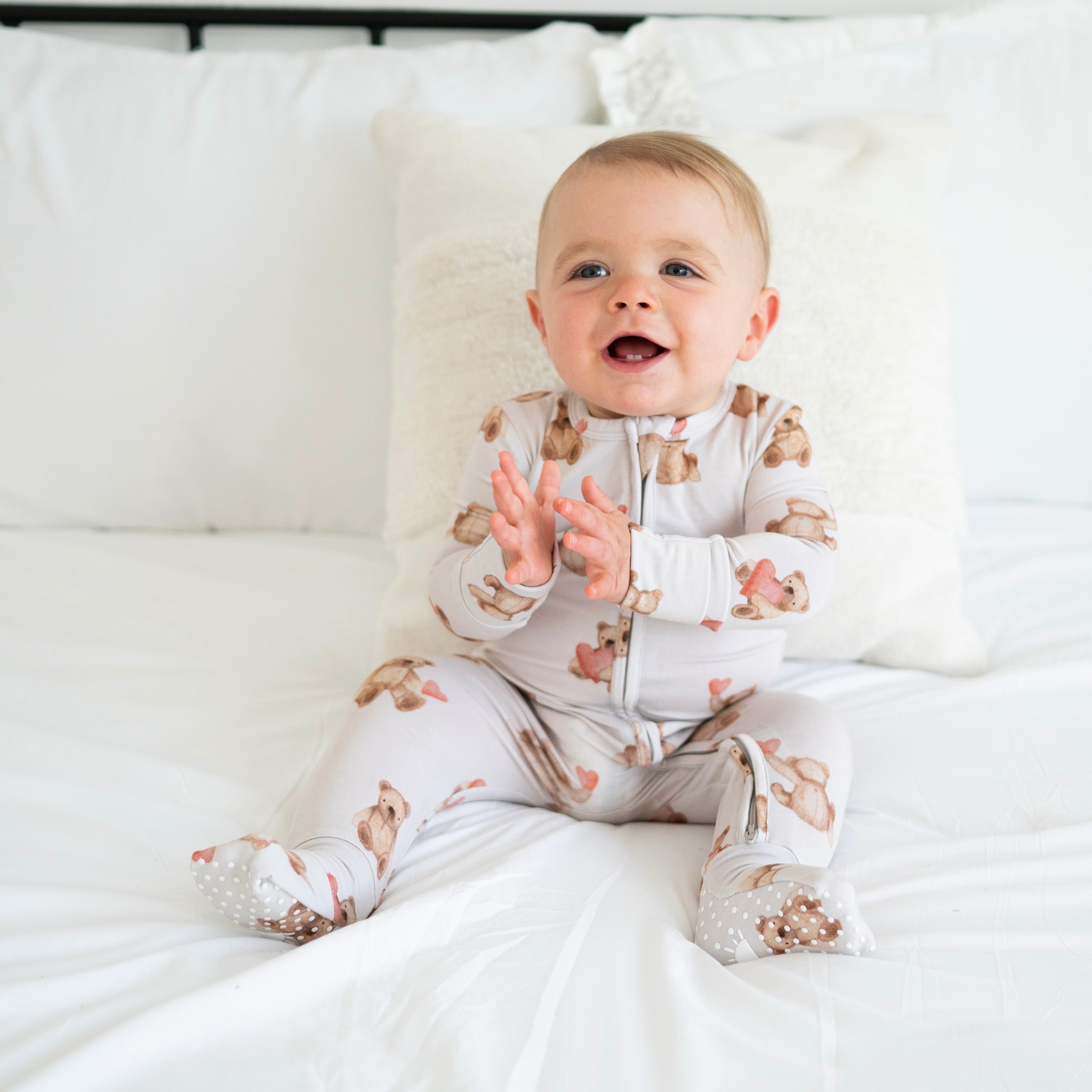 A baby sits on a white bed wearing a light gray zippered pajama set with a teddy bear print.