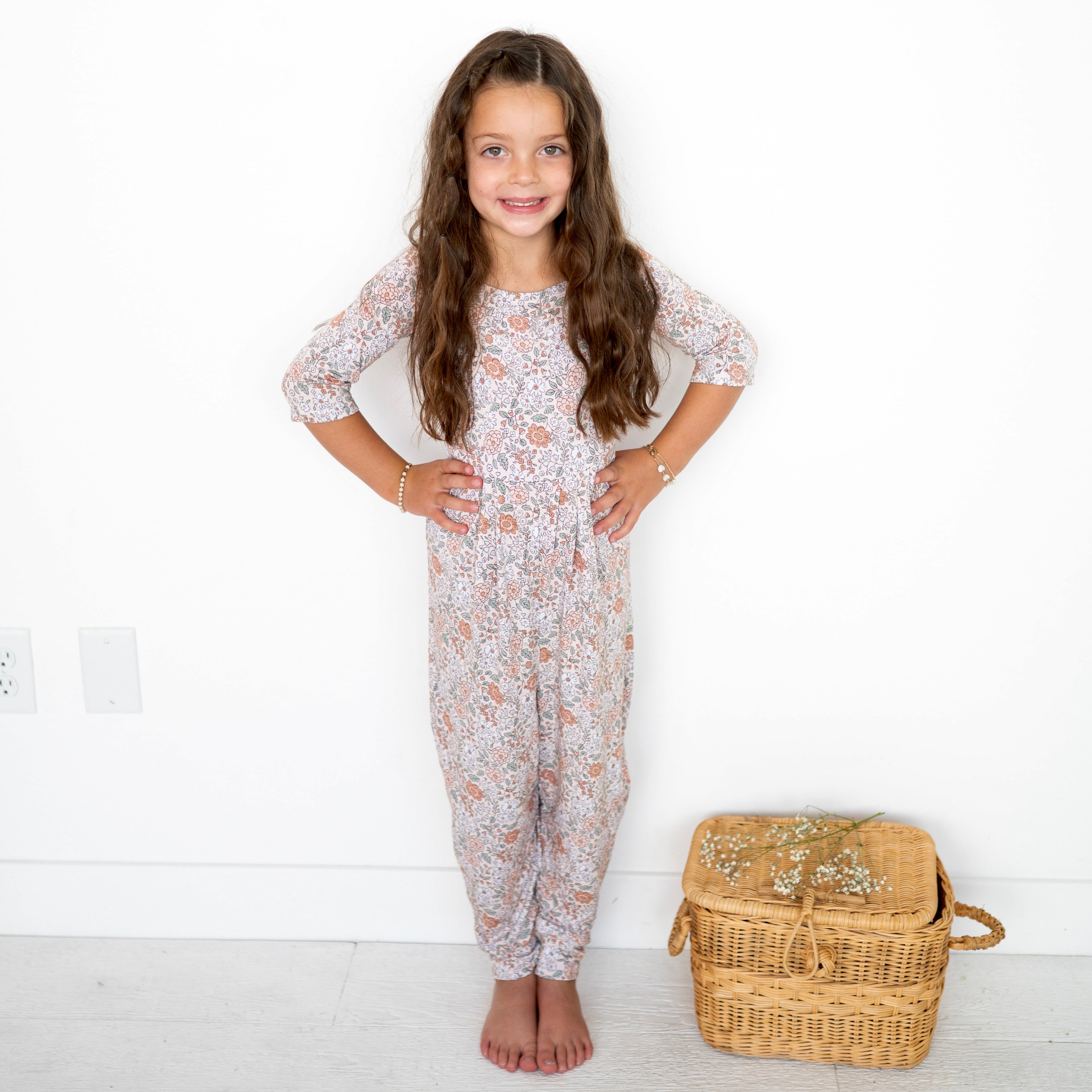 A young girl in a vintage floral romper stands with her hands on her hips next to a wicker basket.