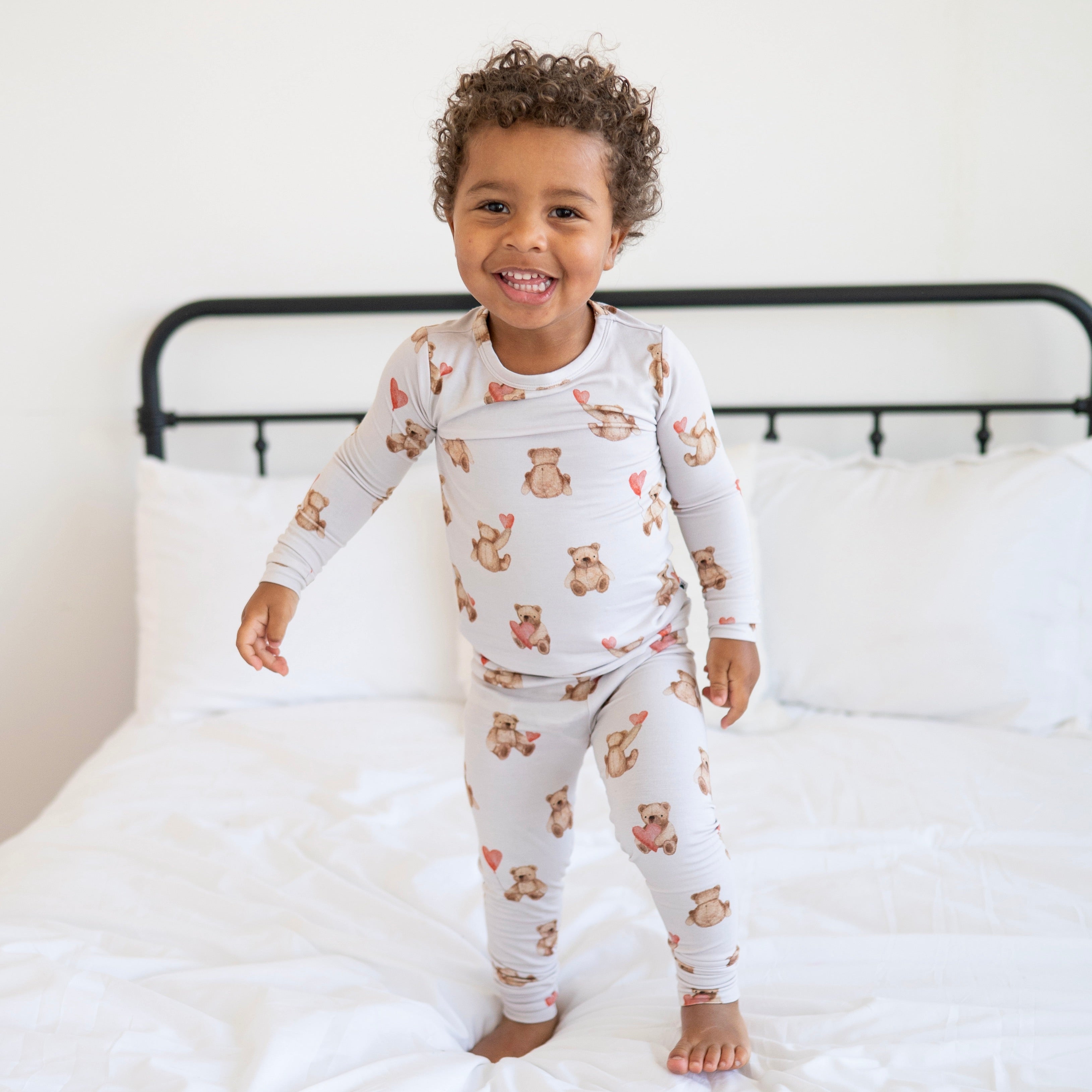 A young child smiles while wearing a light gray pajama set with a teddy bear and heart pattern.
