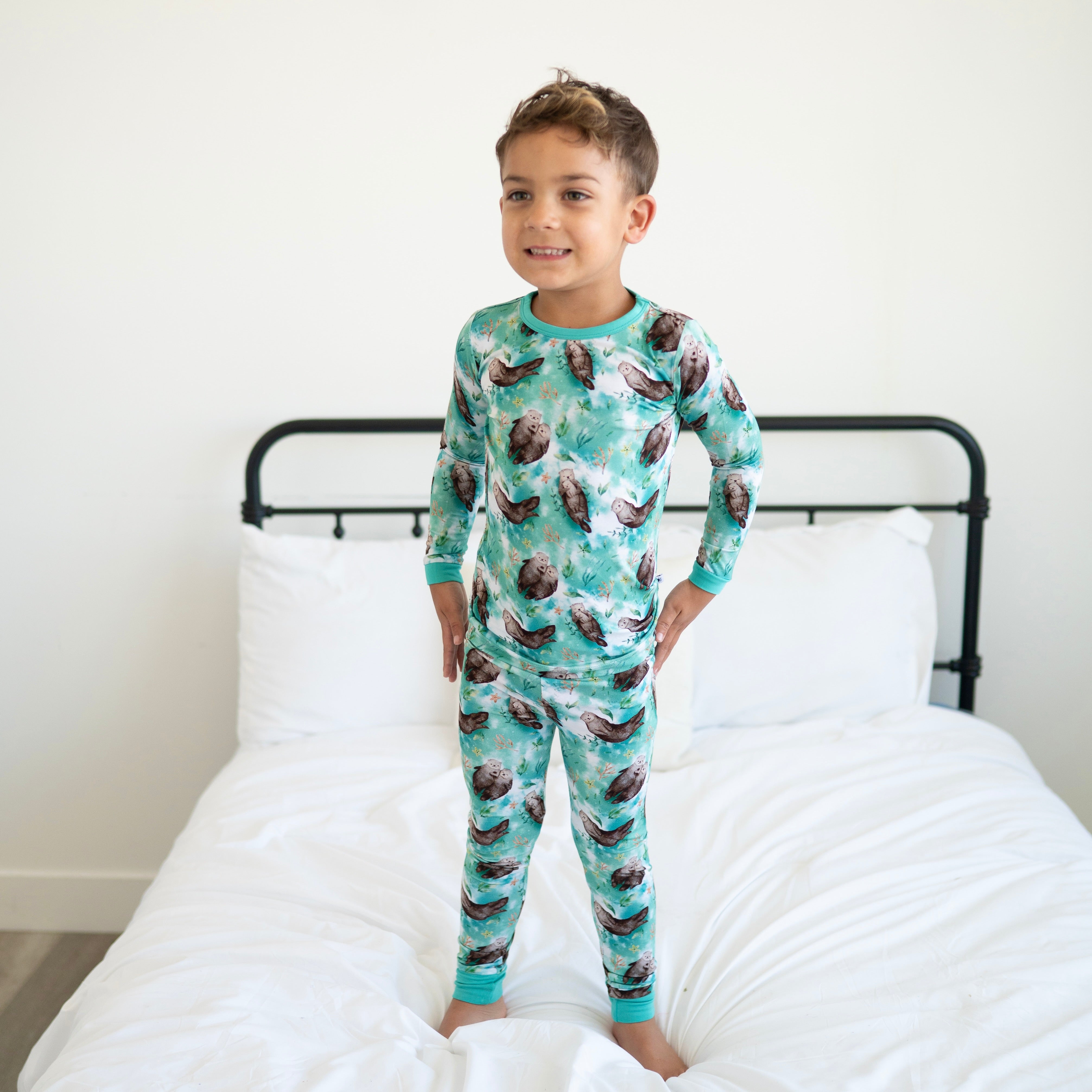 A young boy stands on a bed wearing a long-sleeved pajama set with an otter print.