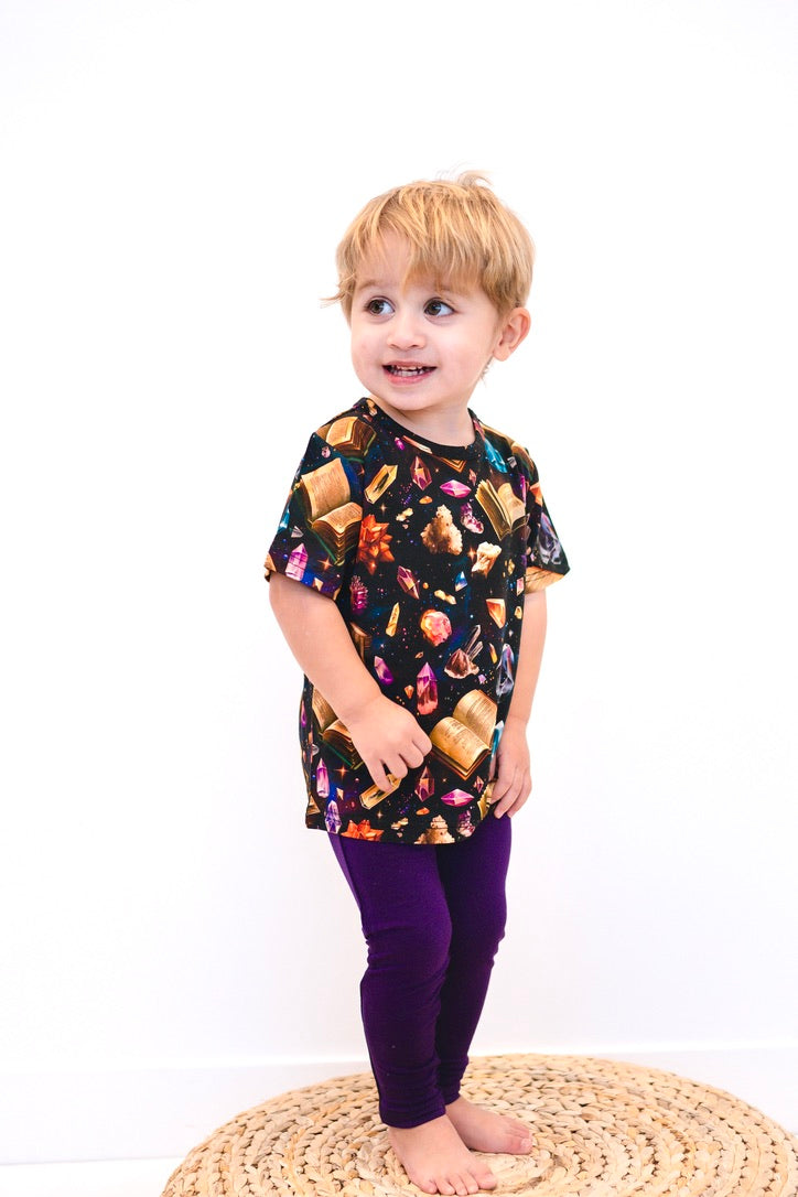 A young child wears a black t-shirt with a crystal and book pattern and purple leggings.