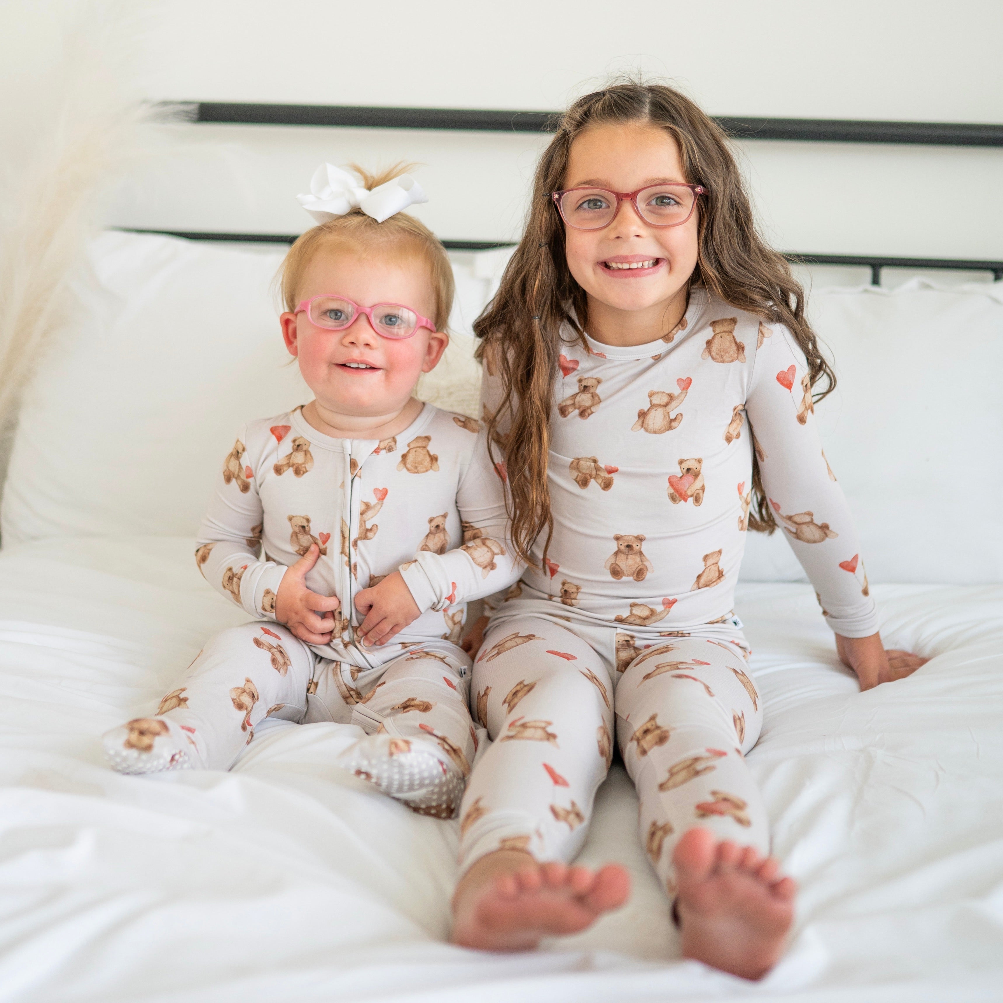 Two children in matching teddy bear print pajamas sit on a bed.