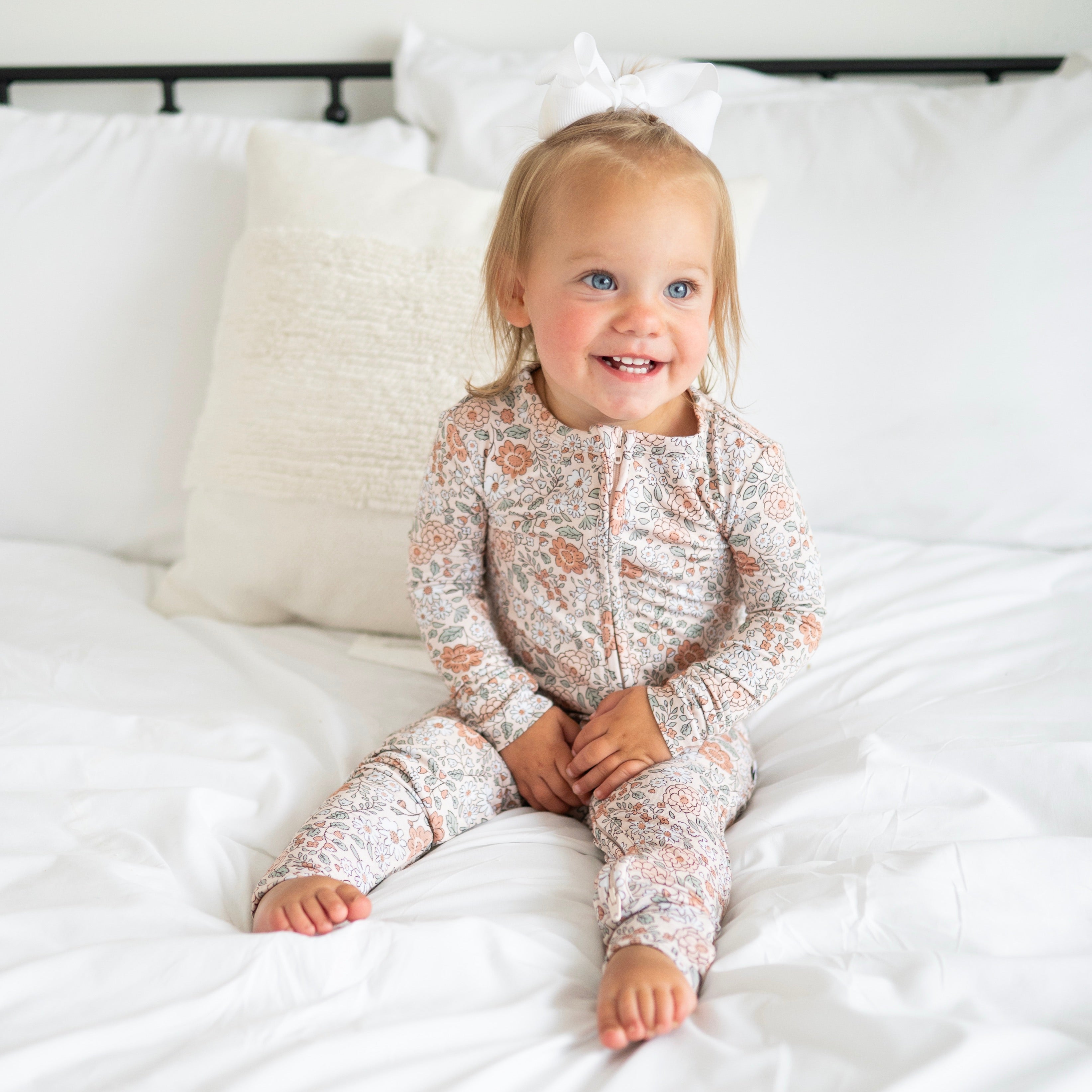A toddler girl in a vintage floral zip-up pajama set sits on a white bed.
