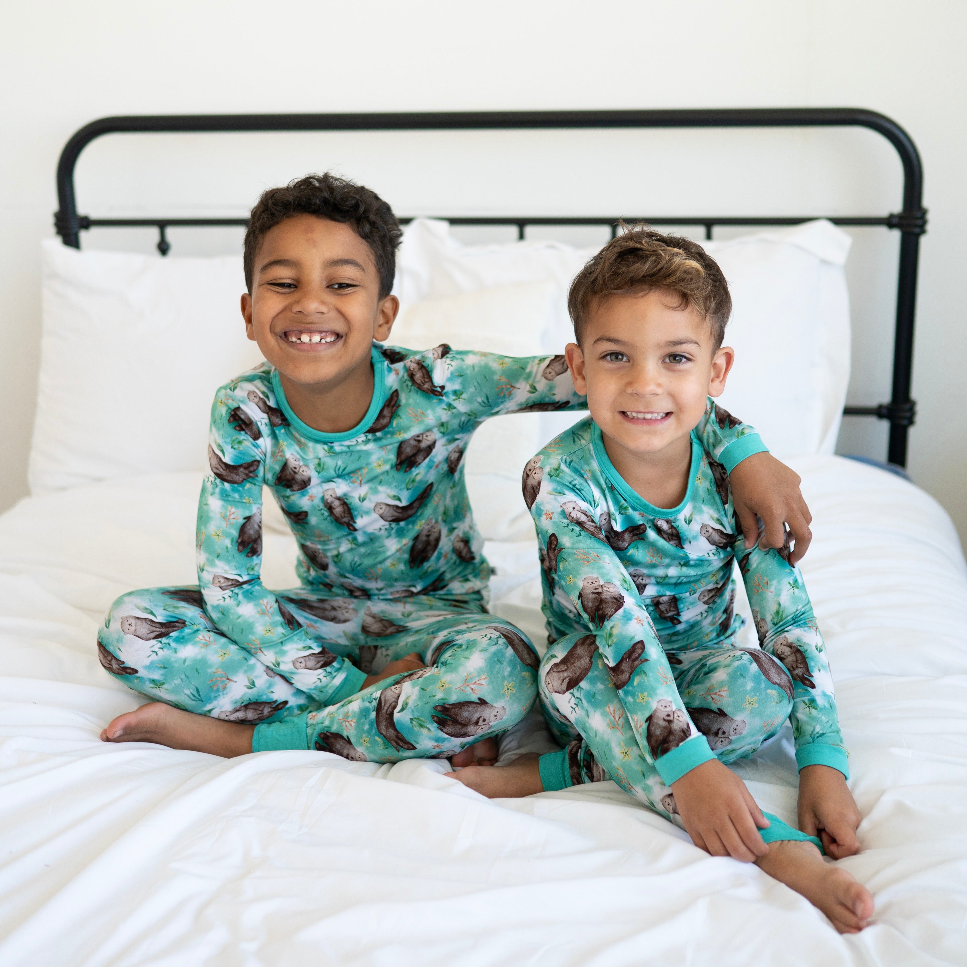 Two boys in teal pajamas with an otter print sit on a white bed.