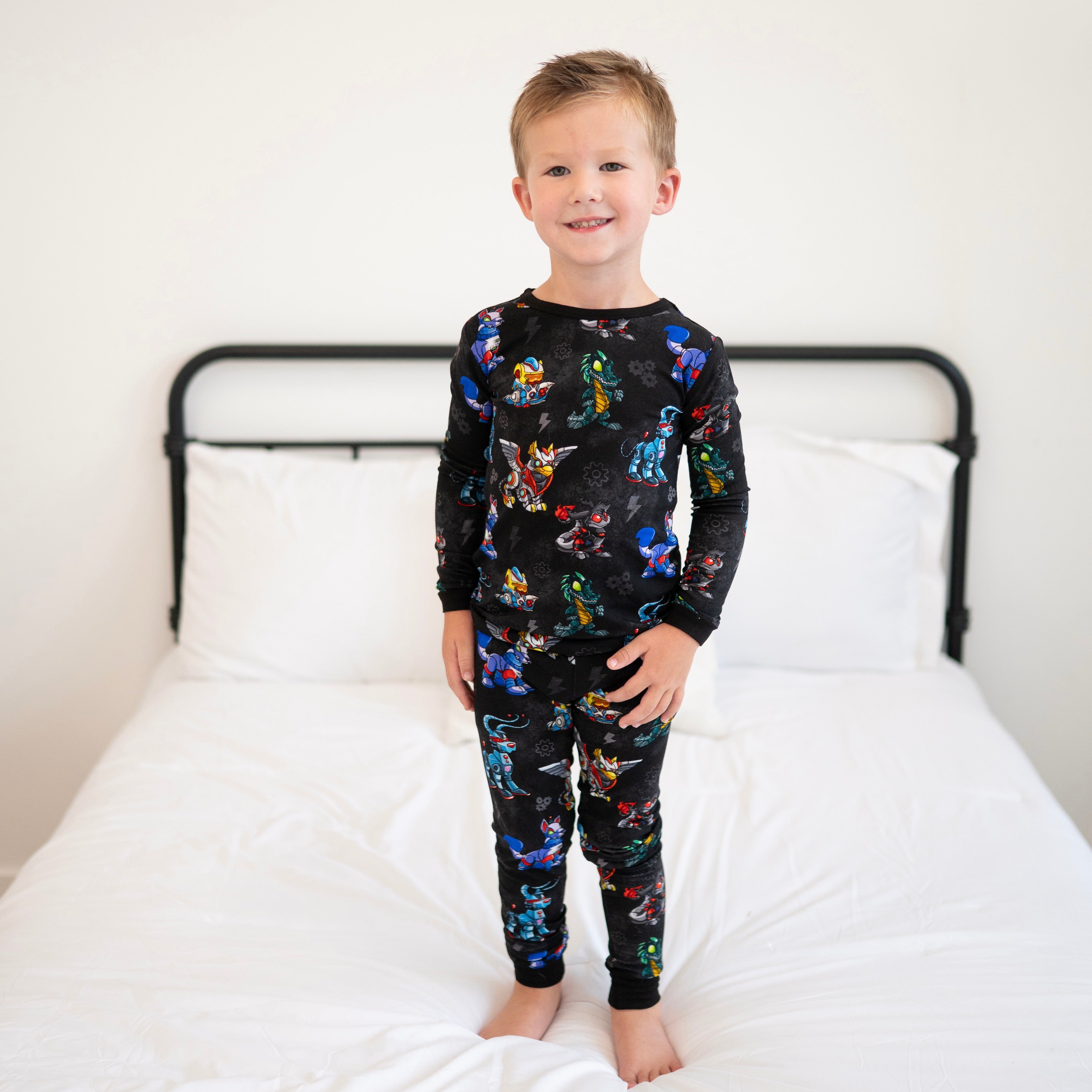 A young boy stands on a white bed wearing black pajamas with a Neopets robot print.