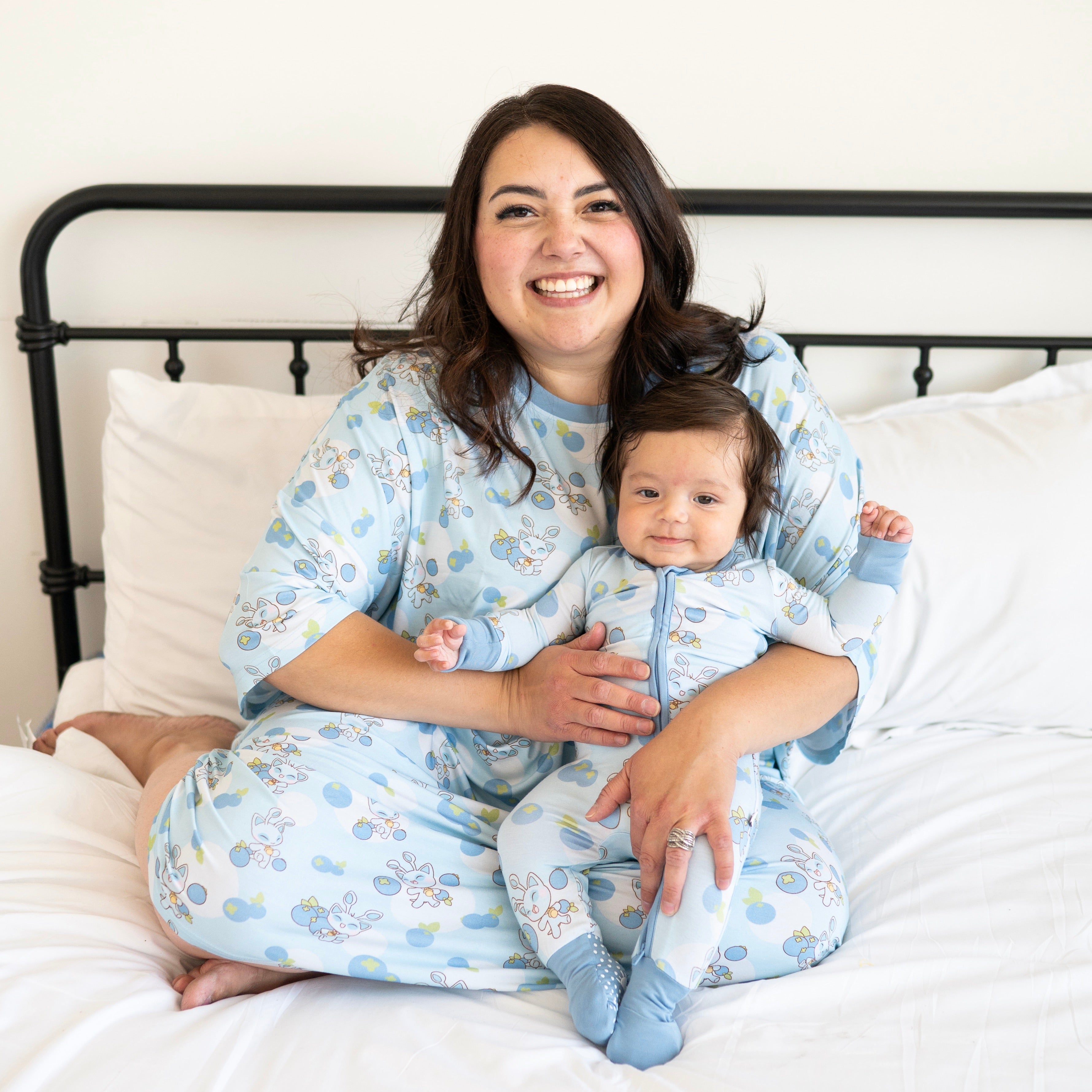 A woman and baby in matching Neopets blueberry pajamas sit on a bed.