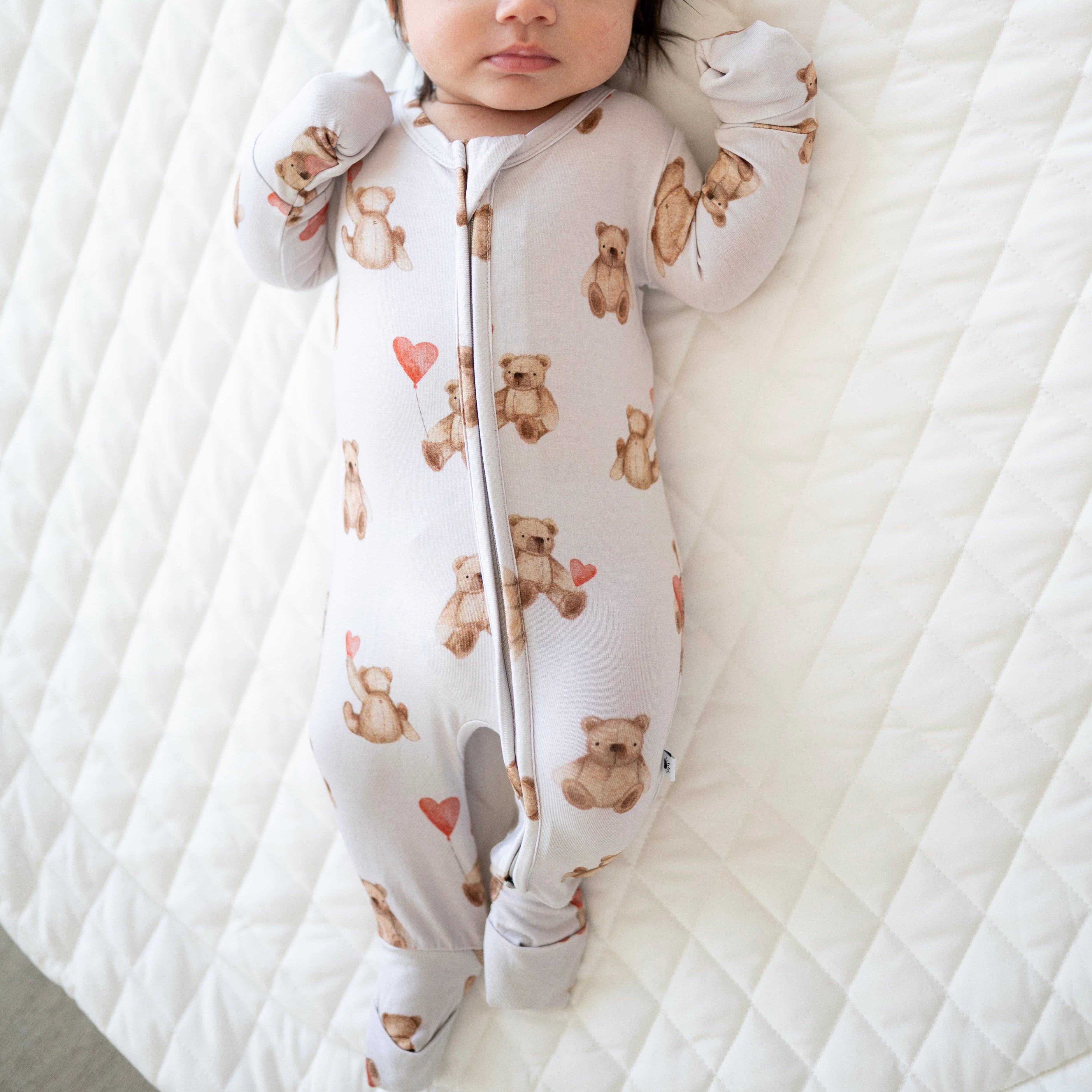 A baby wearing a light grey zippered pajama set with a teddy bear print.