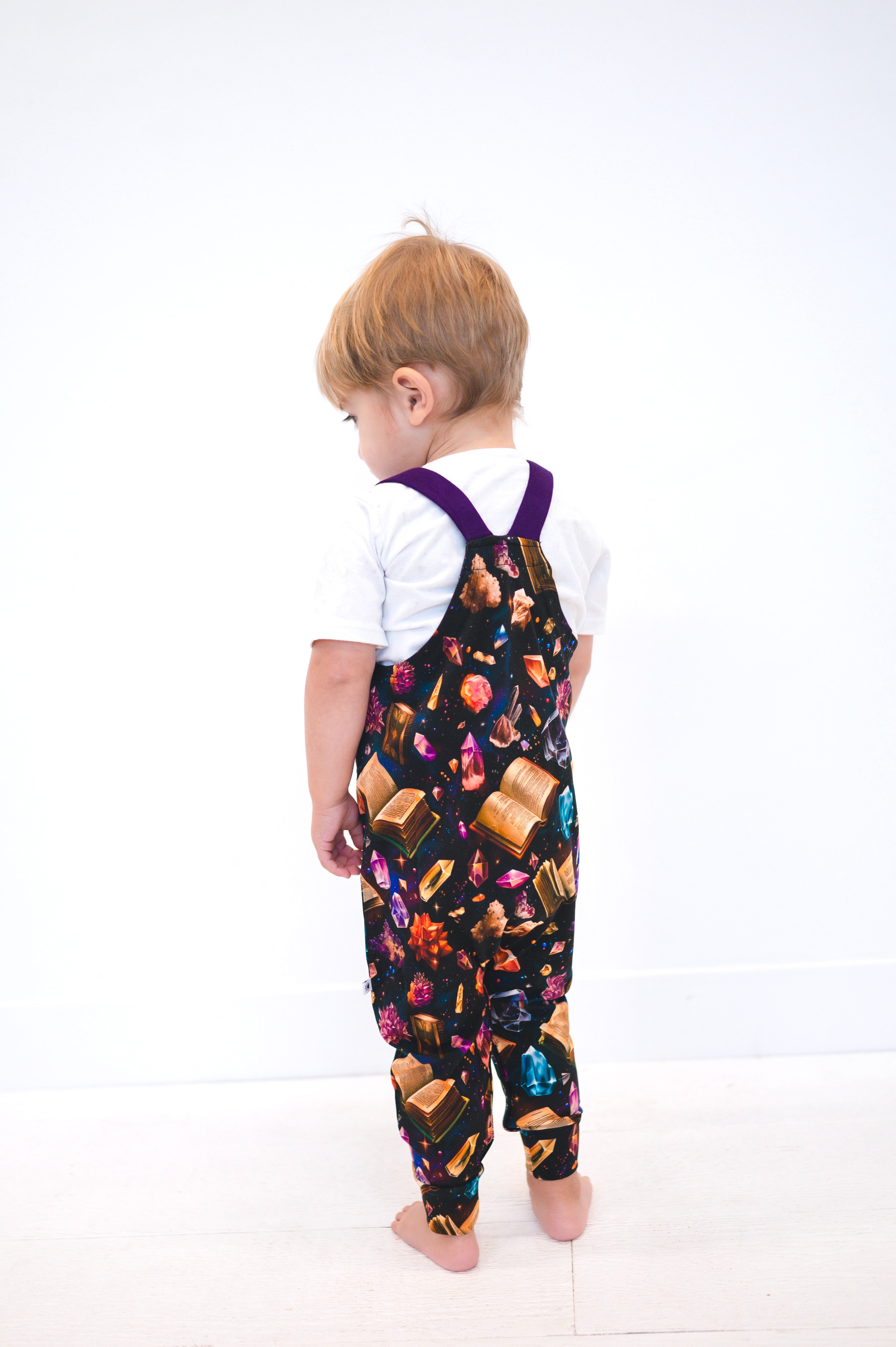 Toddler wearing black overalls with a crystal and book print and purple straps.
