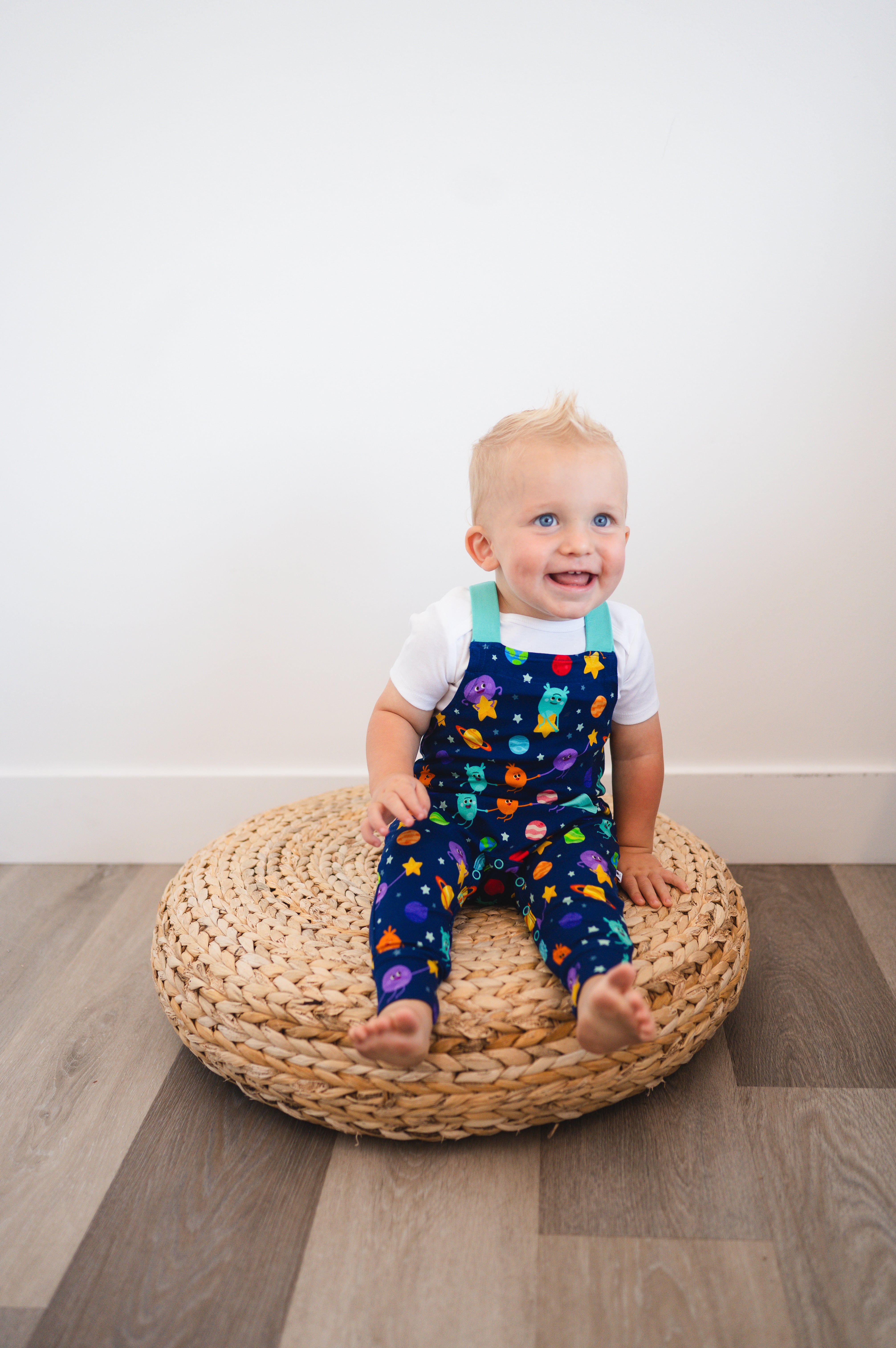 A baby boy wearing blue overalls with a space-themed print sits on a woven cushion.