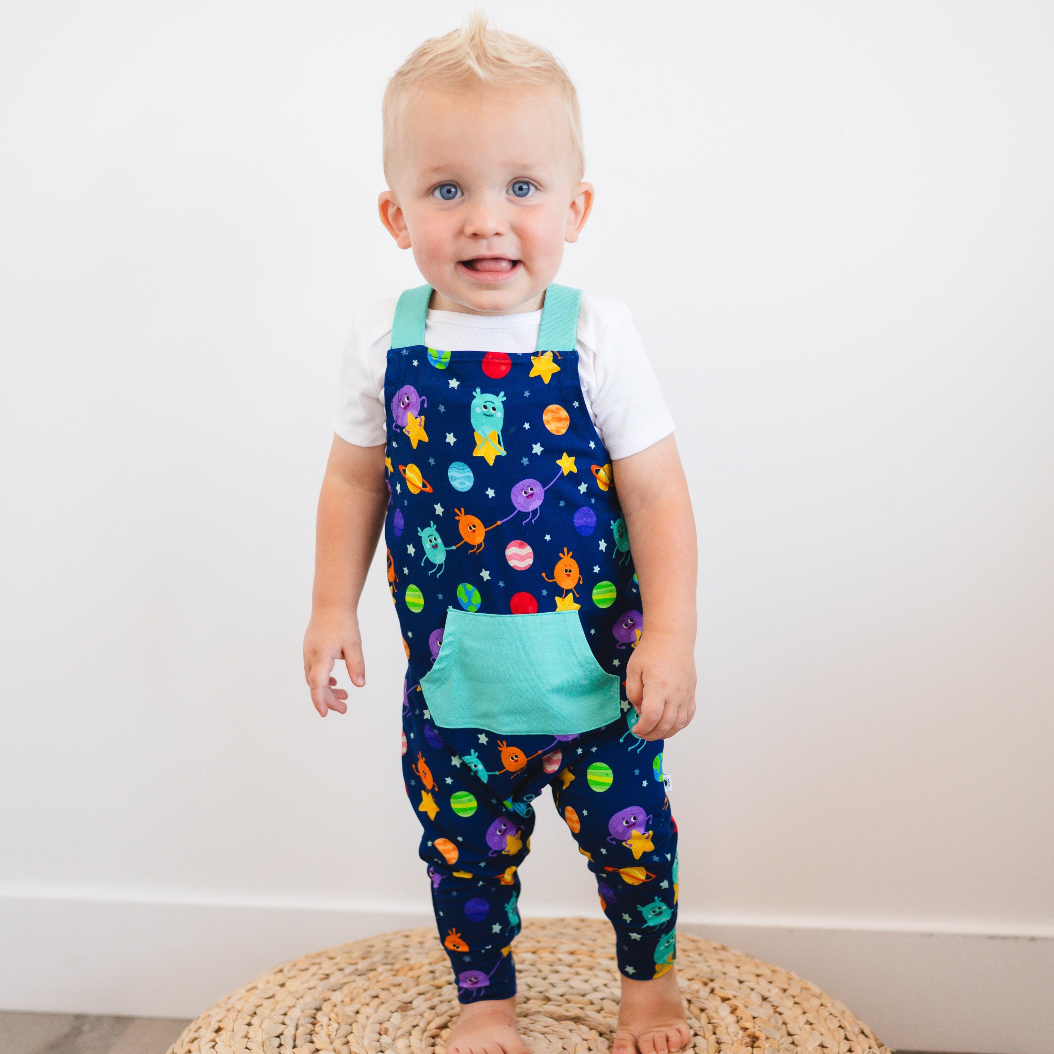 A toddler boy wears navy blue overalls with a space-themed print and a teal pocket.