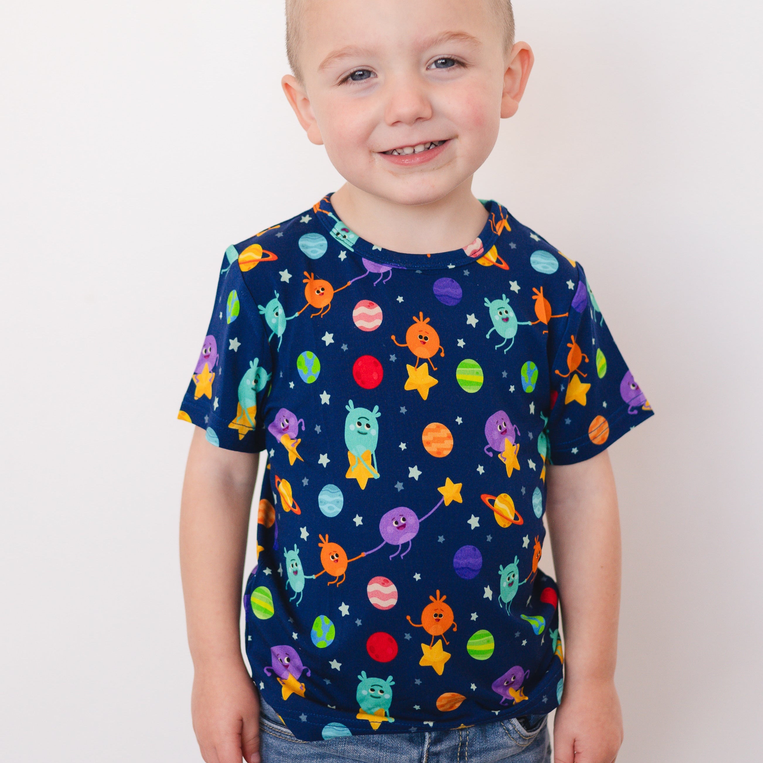 A young boy smiles wearing a navy blue t-shirt with a colorful space-themed print.