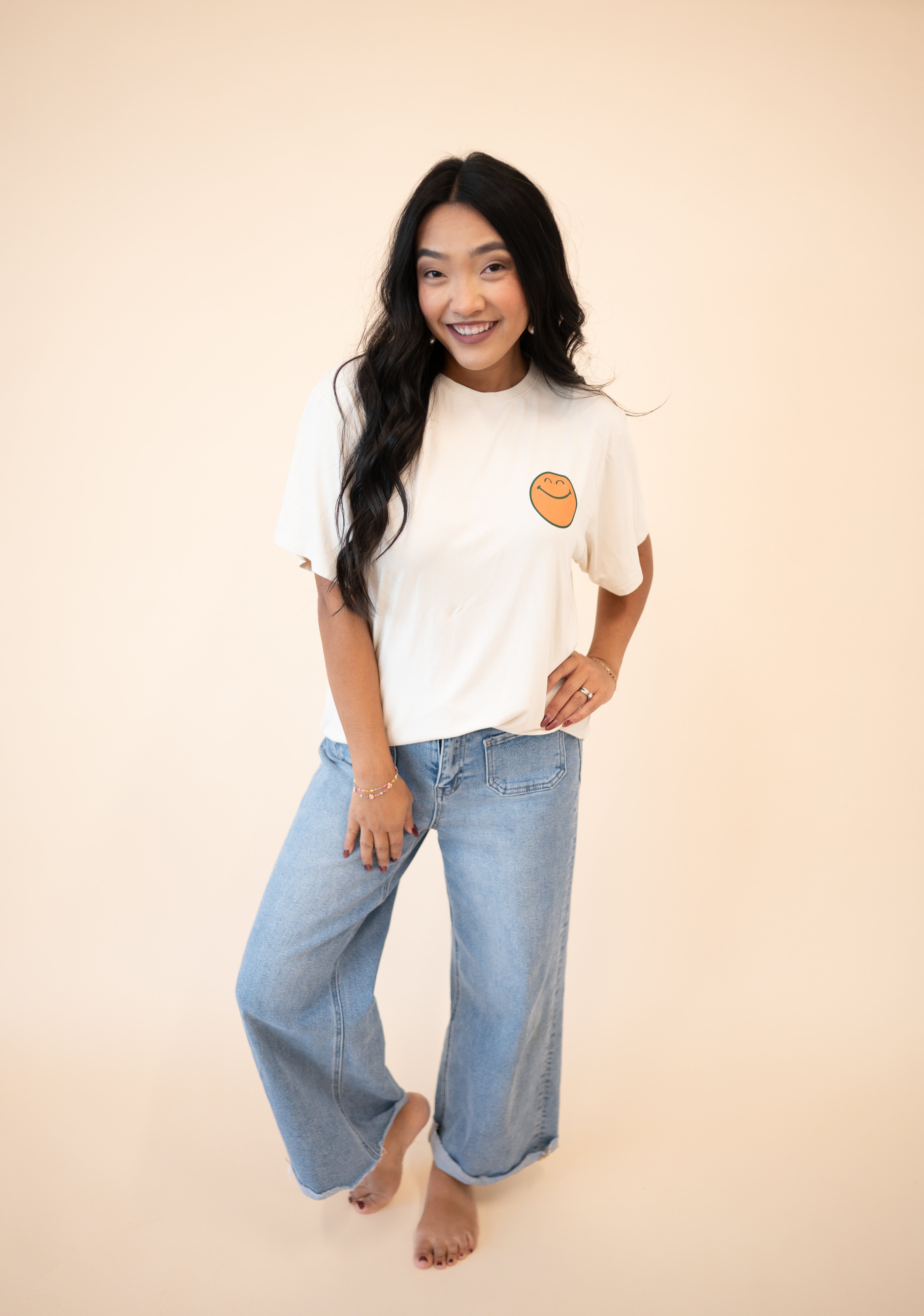 A woman wears an oversized cream t-shirt with a smiley face graphic and light wash wide-leg jeans.
