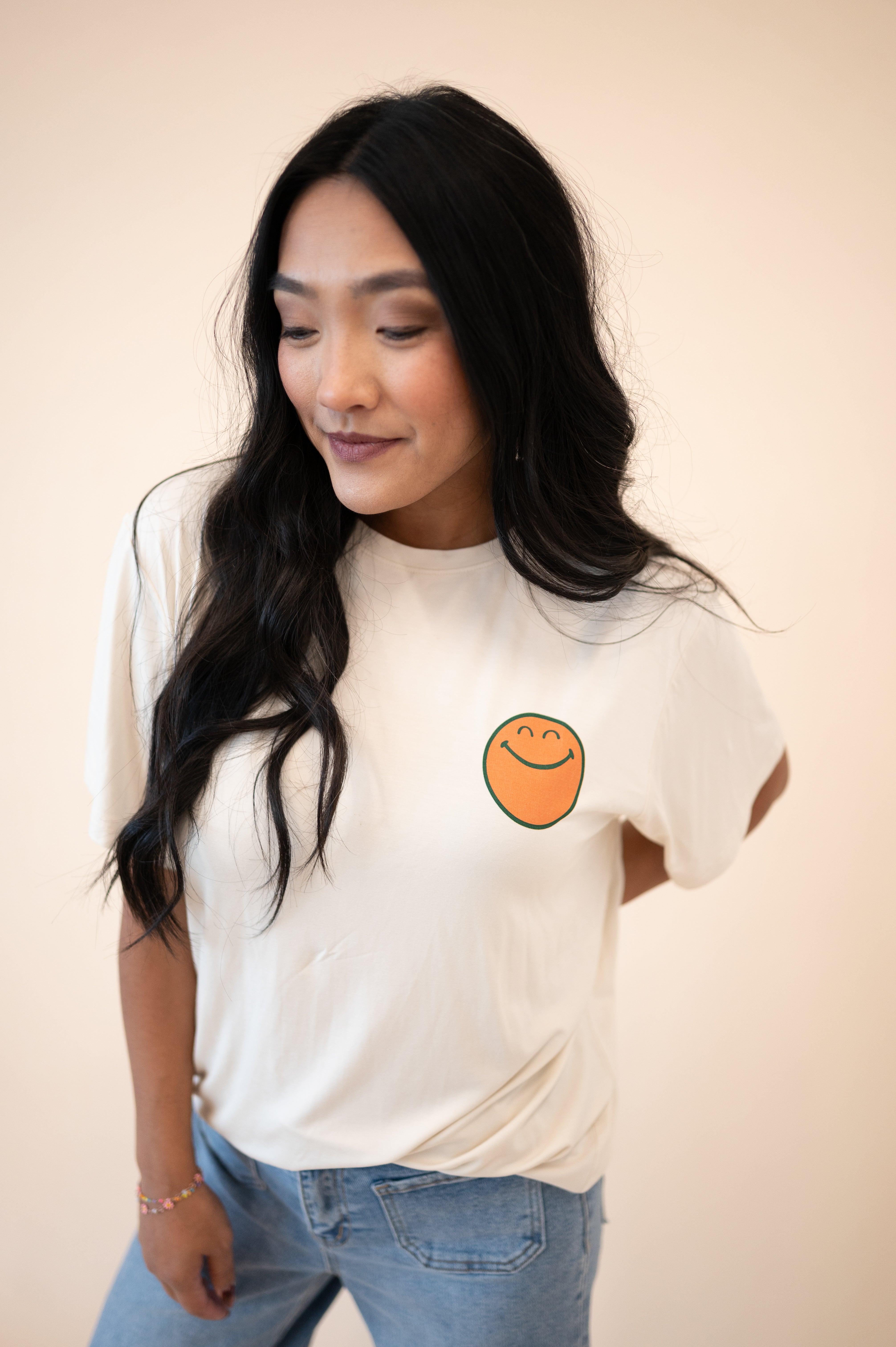 A woman wears an oversized cream t-shirt with a smiley face graphic and blue jeans.