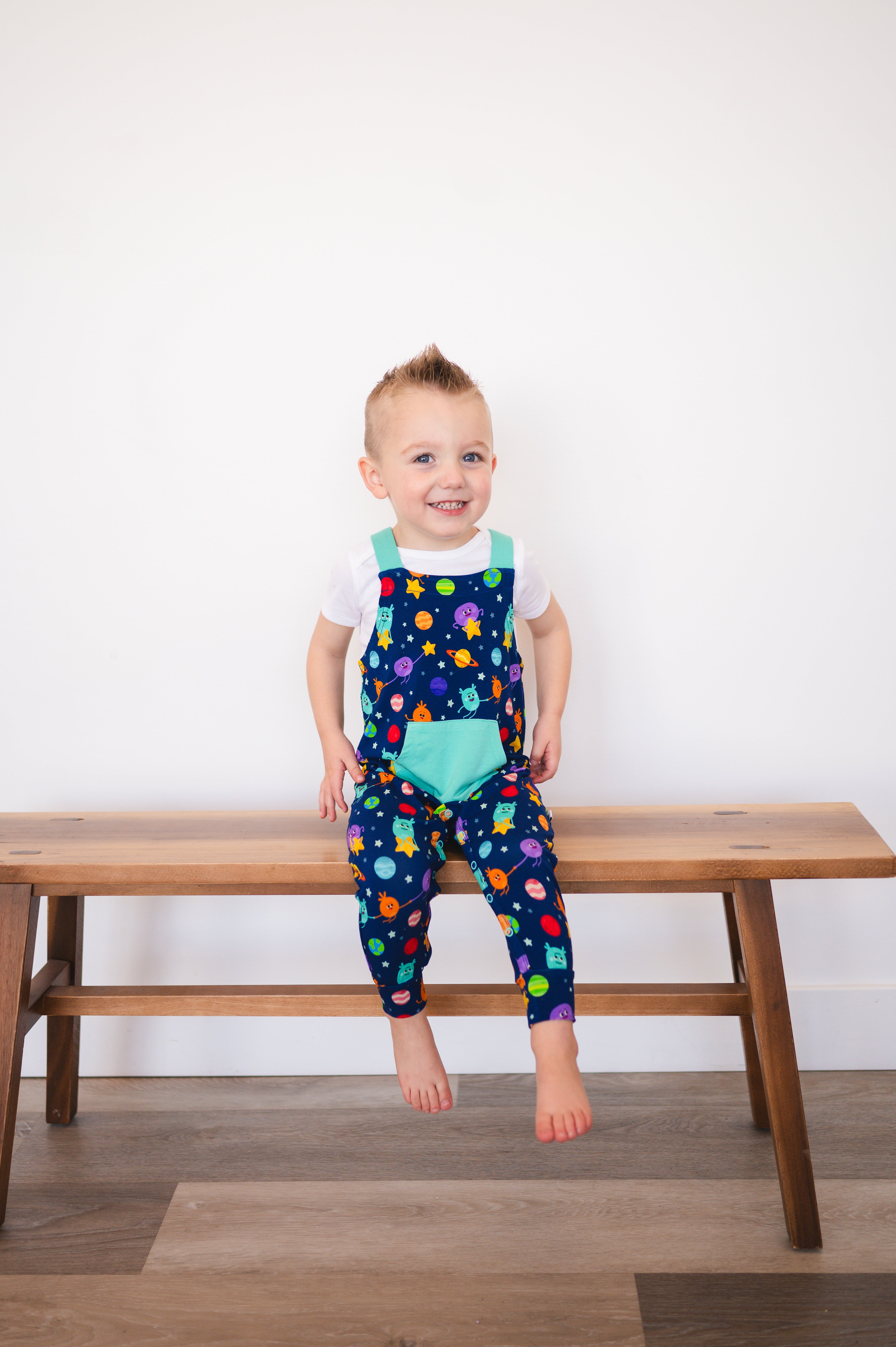 A young boy wears blue overalls with a space-themed print, sitting on a wooden bench.