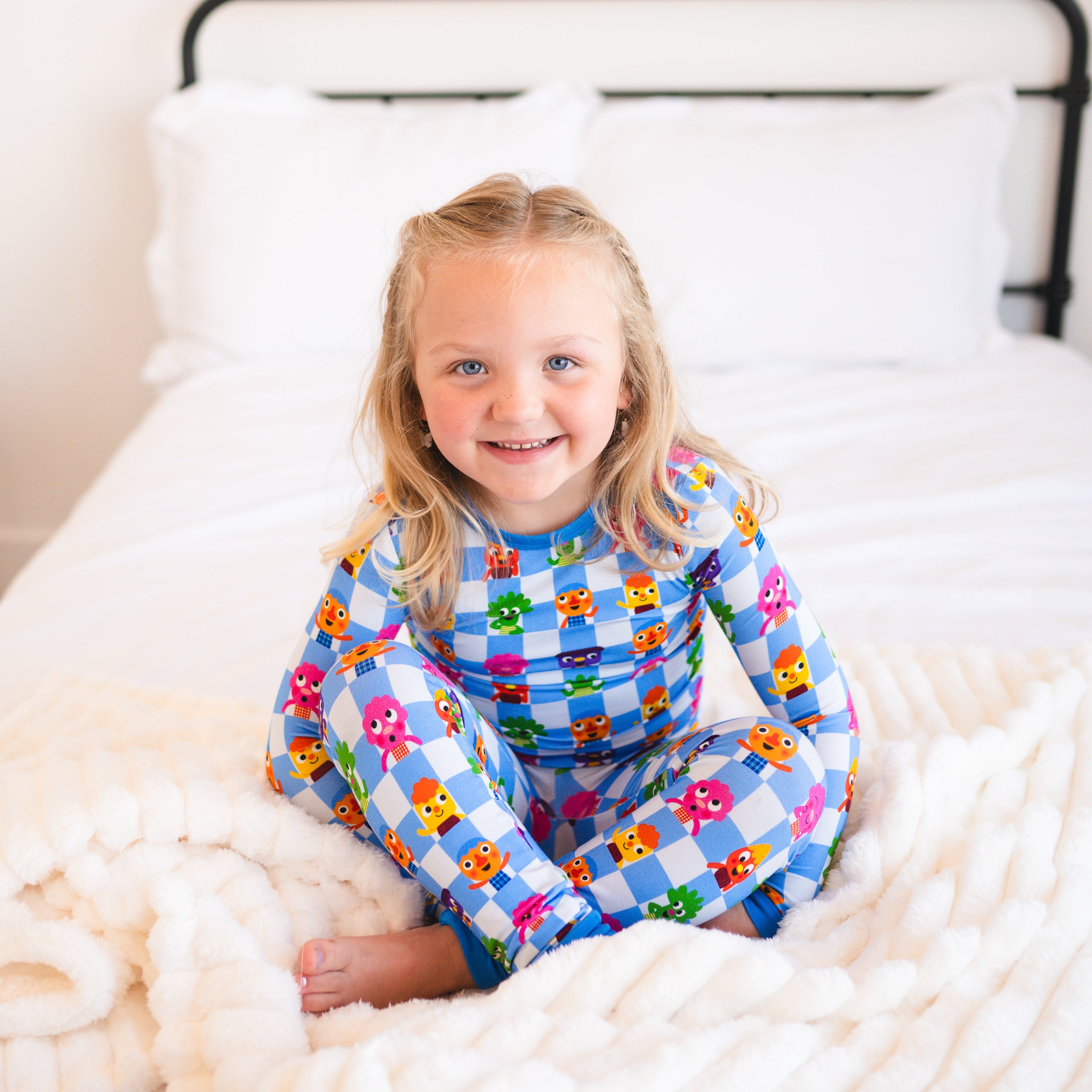 A young girl in blue and white checkered pajamas with cartoon characters sits on a fluffy white blanket.