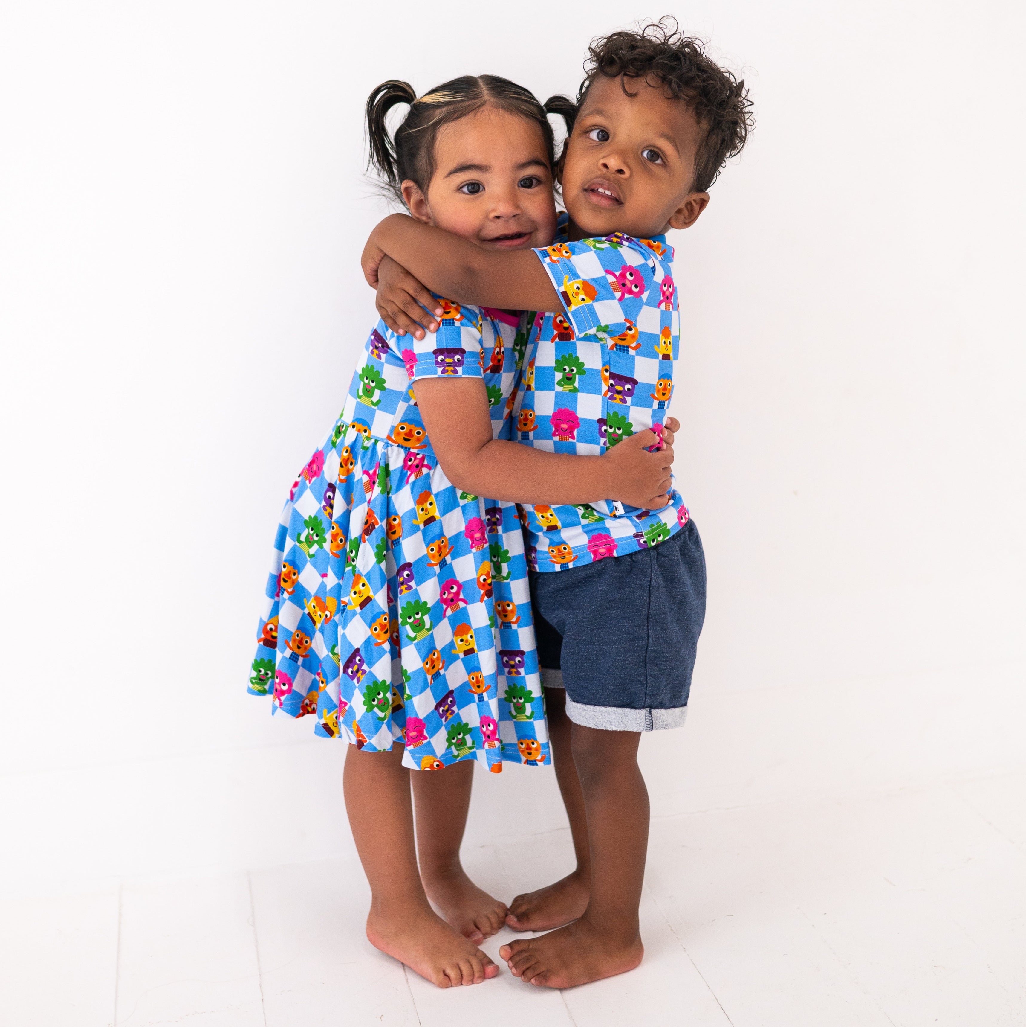 A girl in a blue checkered dress with cartoon characters hugs a boy in a matching shirt and denim shorts.