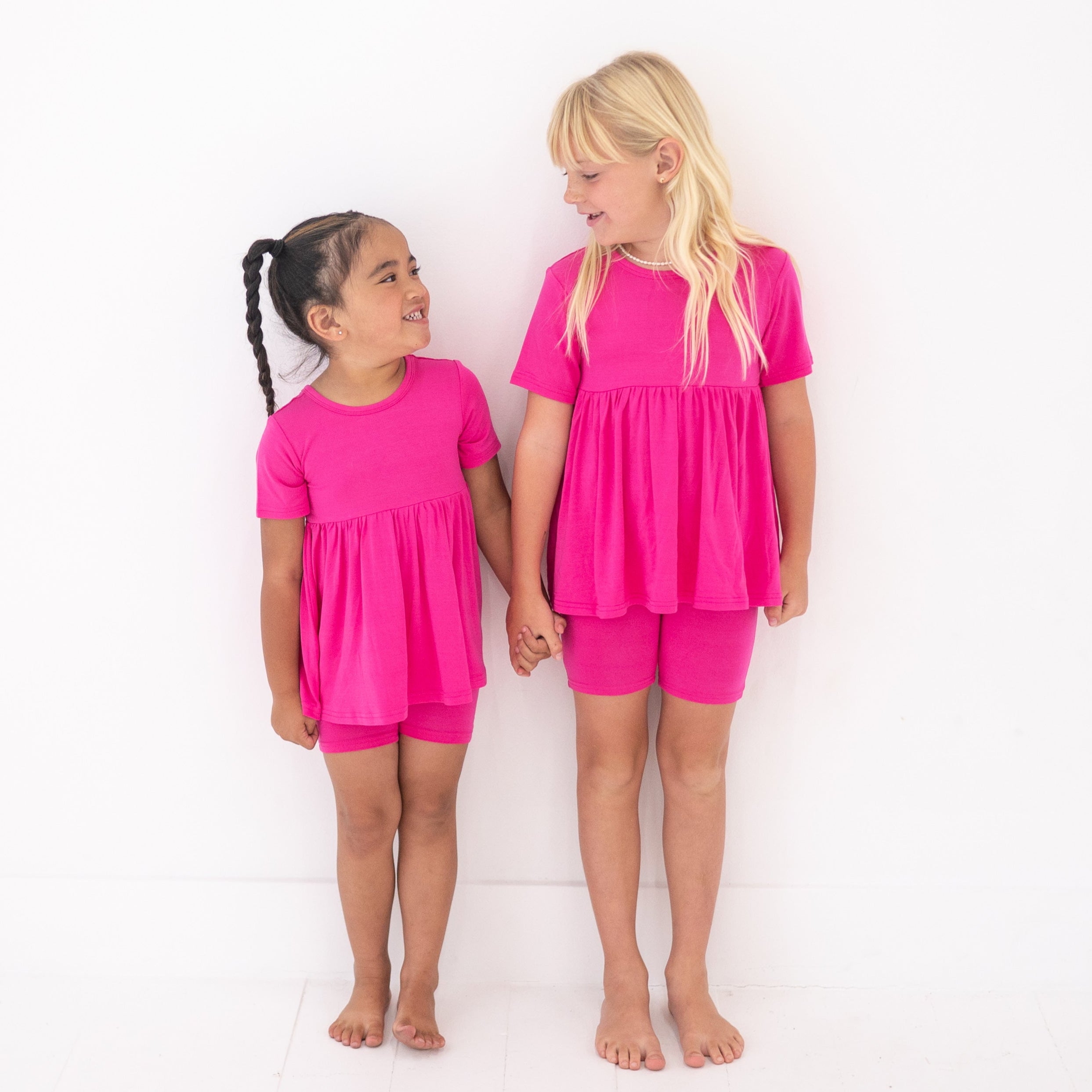 Two young girls in matching pink peplum tops and shorts stand holding hands.