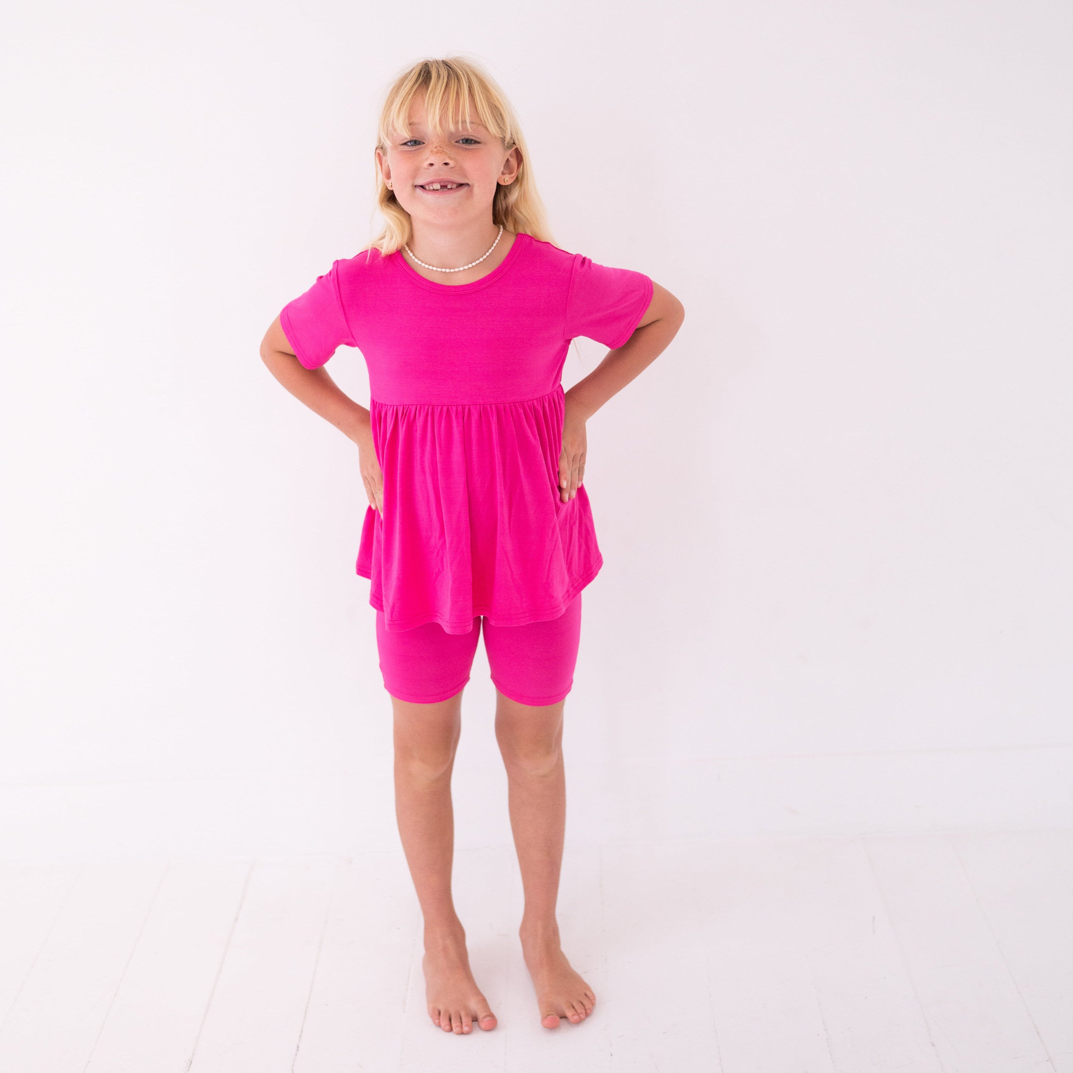 A young girl smiles, wearing a blossom pink peplum top and matching biker shorts.