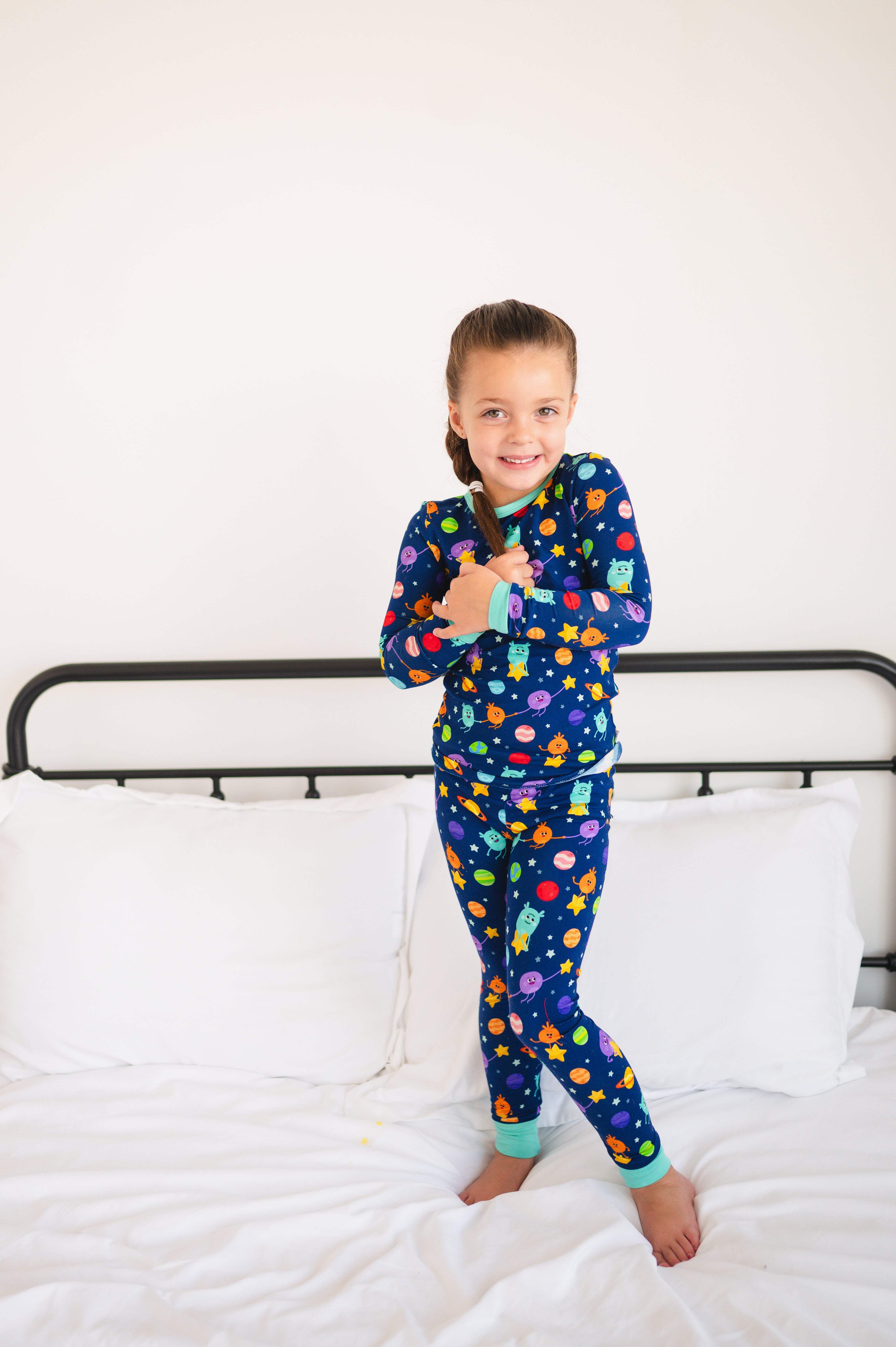 A young girl in blue pajamas with a space-themed print stands on a bed.