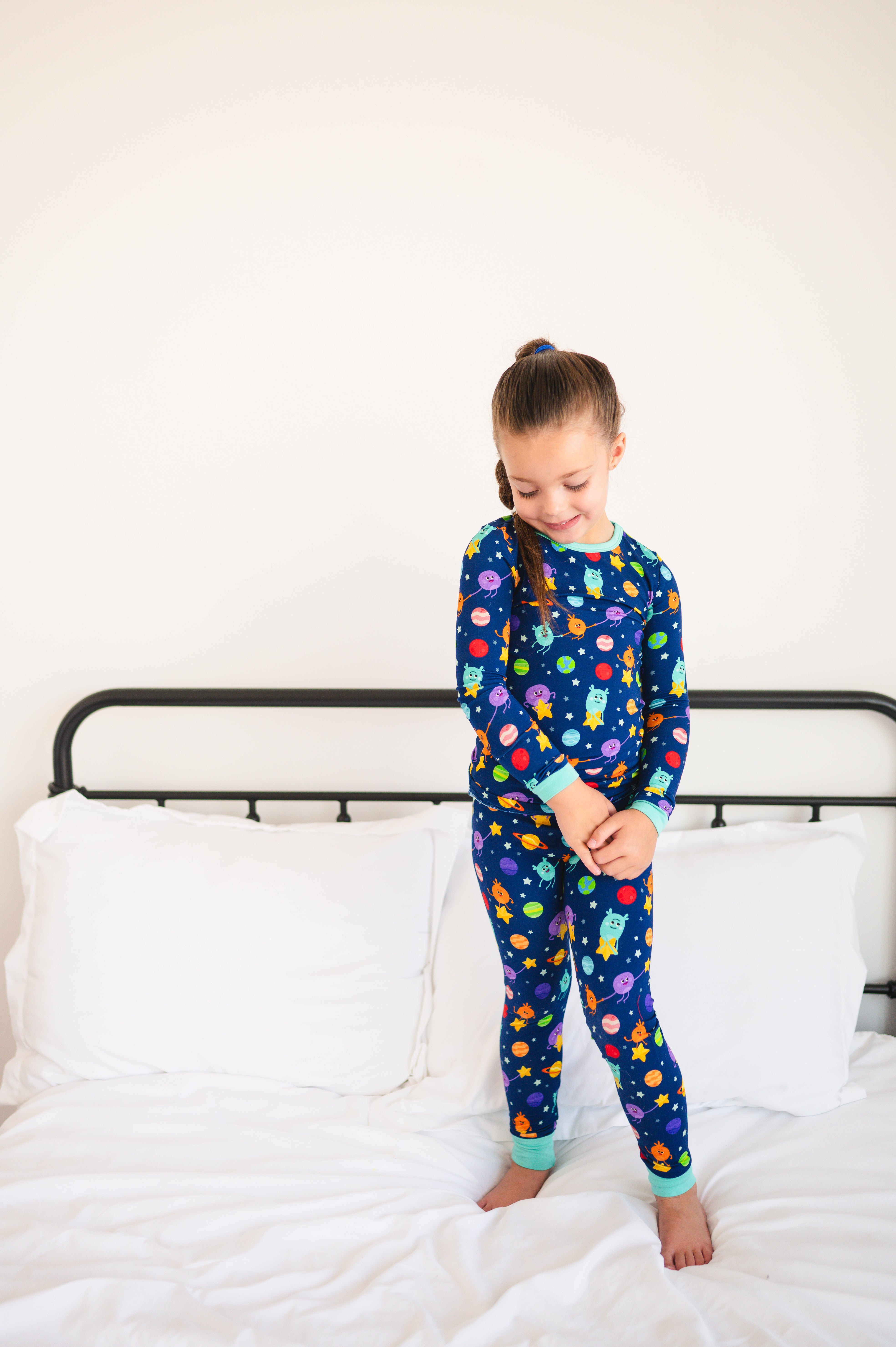 A young girl in blue pajamas with a space-themed print stands on a bed.