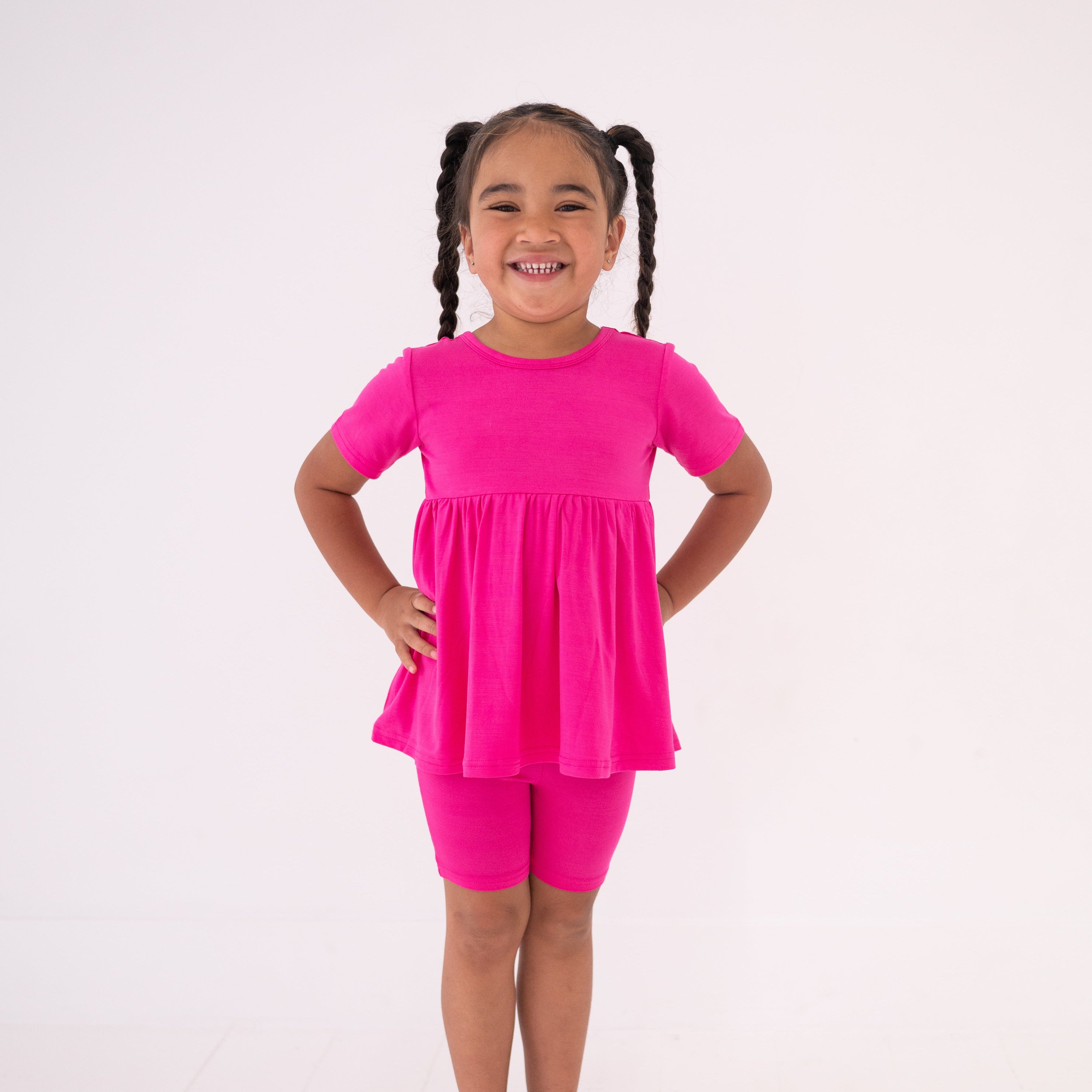 A young girl smiles wearing a pink peplum top and matching biker shorts.