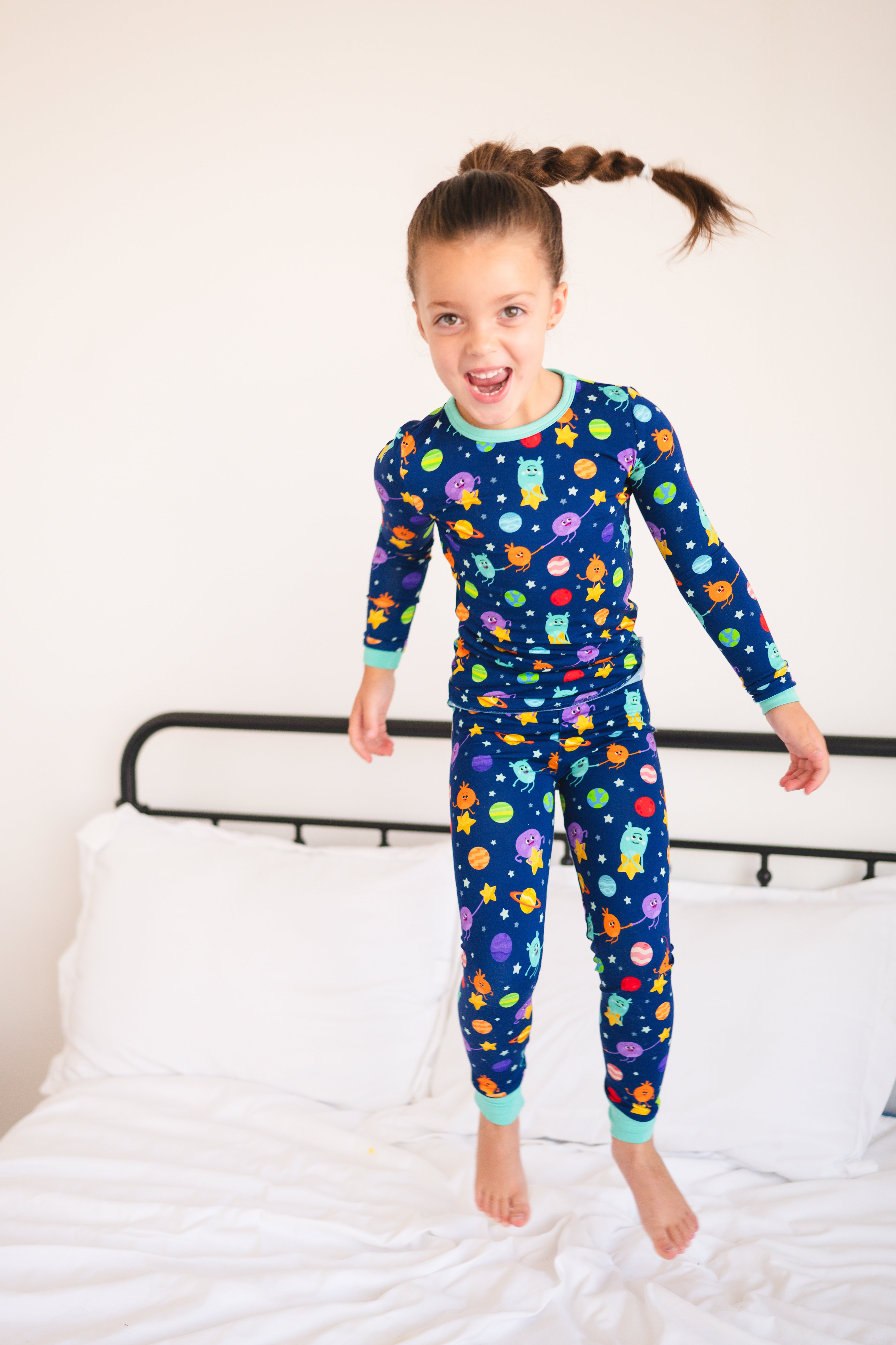 A young girl in blue pajamas with a space-themed print jumps on a bed.