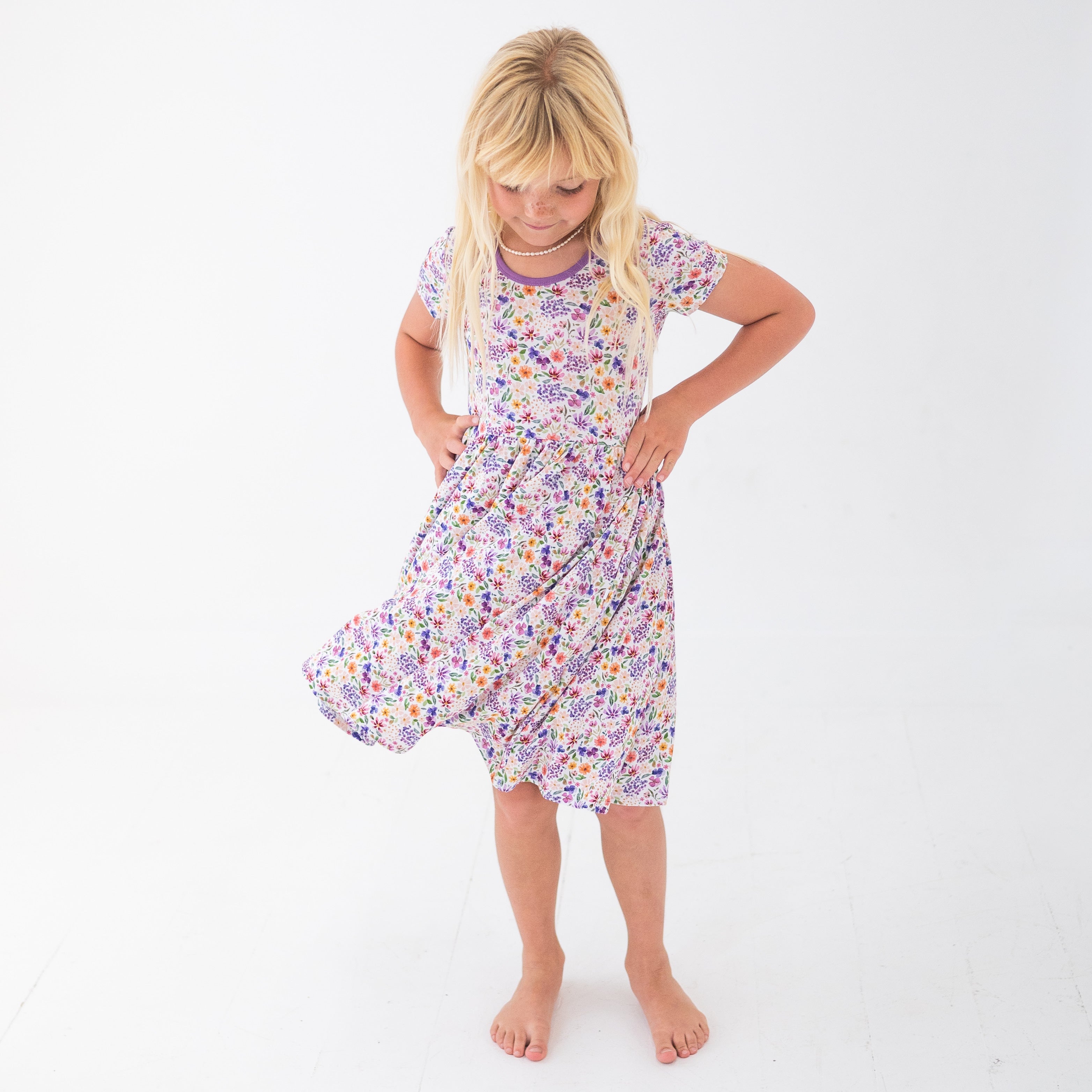 A young girl in a white floral dress with purple trim stands with her hands on her hips.