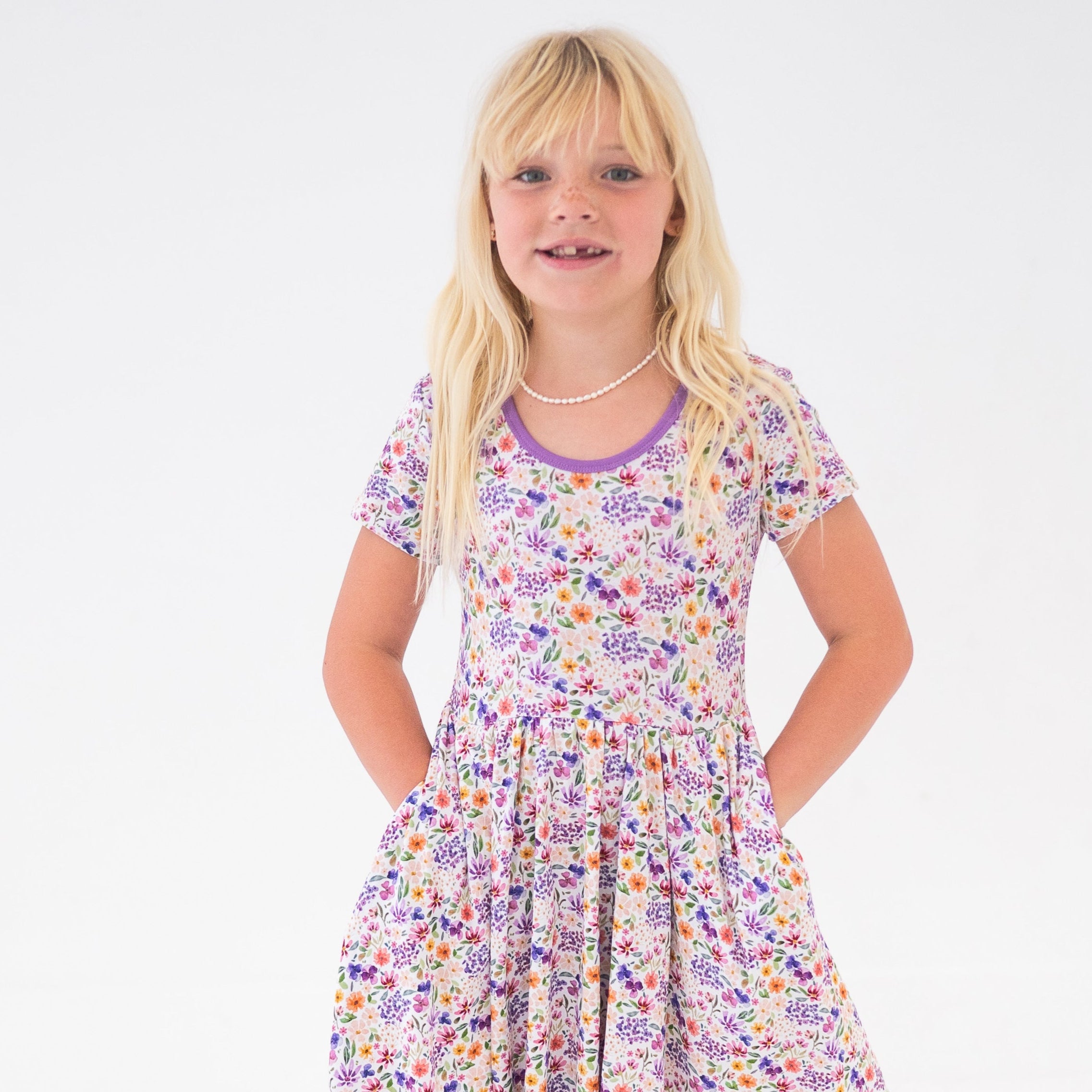 A young girl with blonde hair wears a floral print dress with pockets.