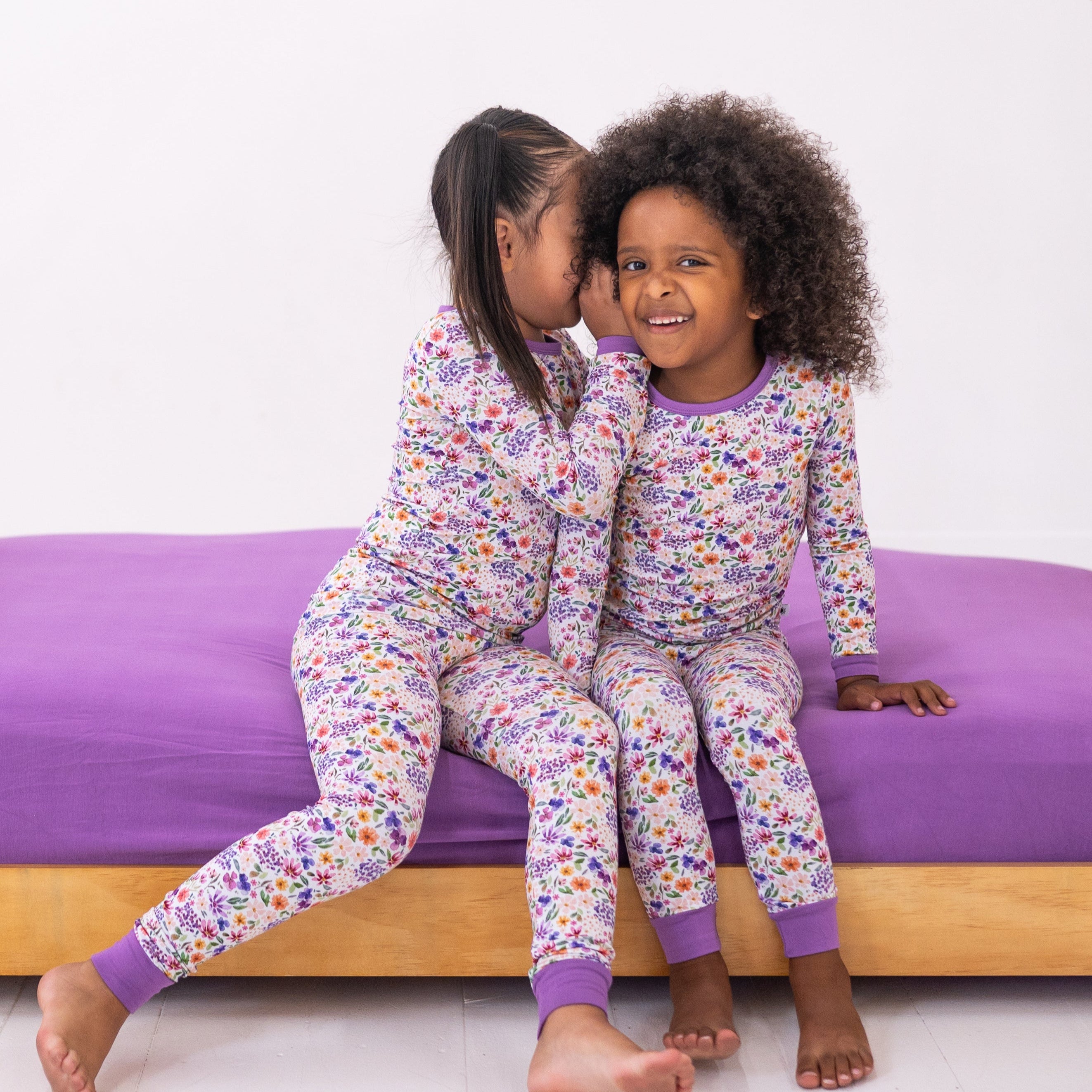 Two girls in floral pajamas sit on a purple bed.