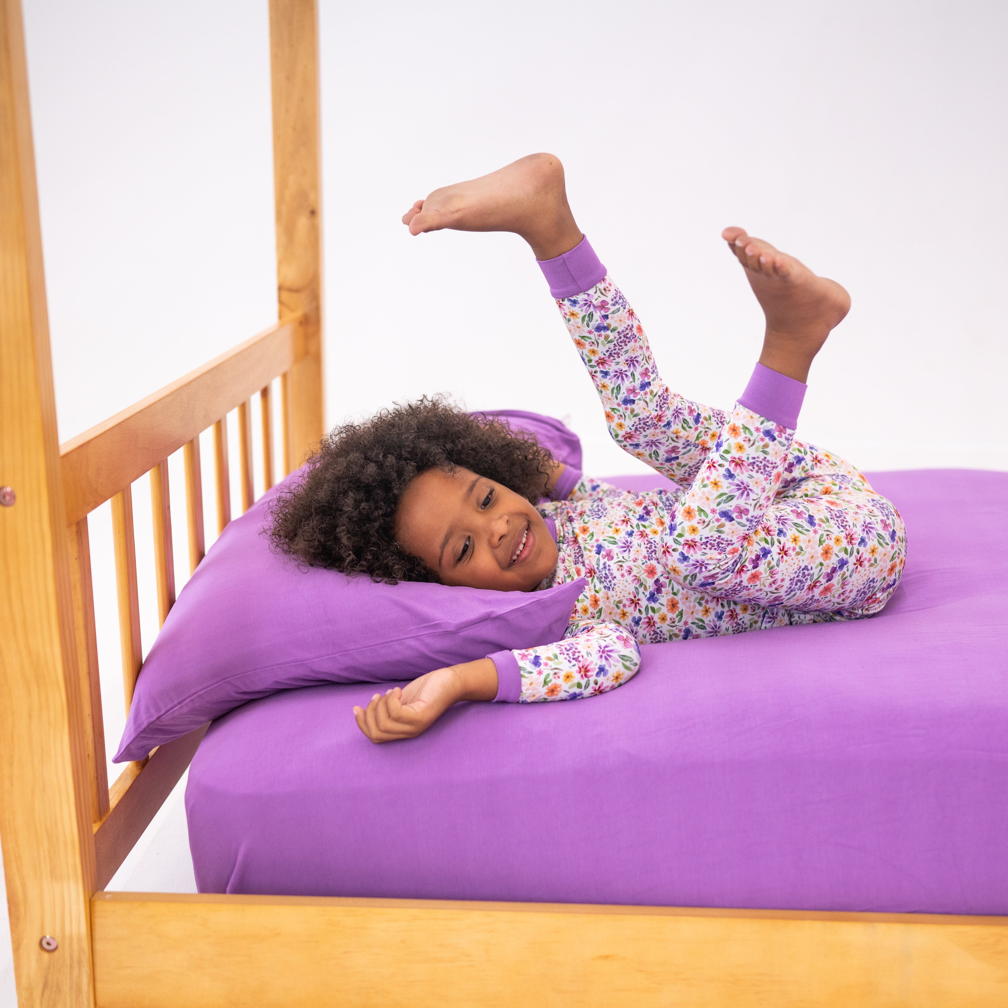 A young girl in floral pajamas plays on a twin bed with lavender sheets.