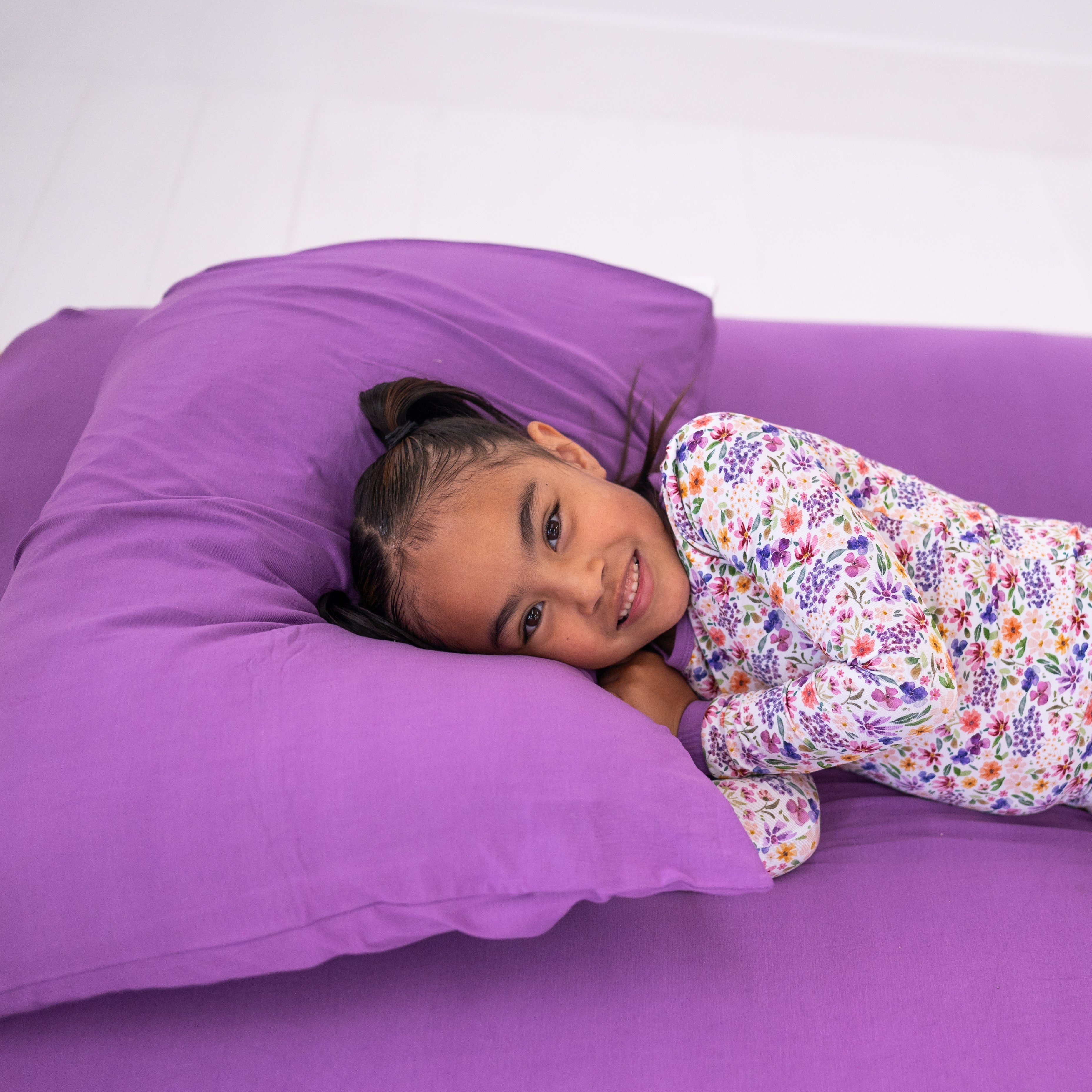 A young girl in a floral pajama set smiles while lying on a lavender fitted twin sheet and pillowcase.